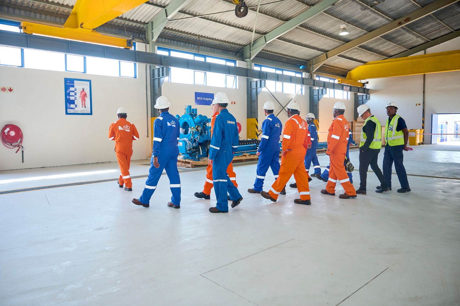 Workers in blue and orange safety coveralls and hard hats walk through an industrial warehouse facility during a site tour.