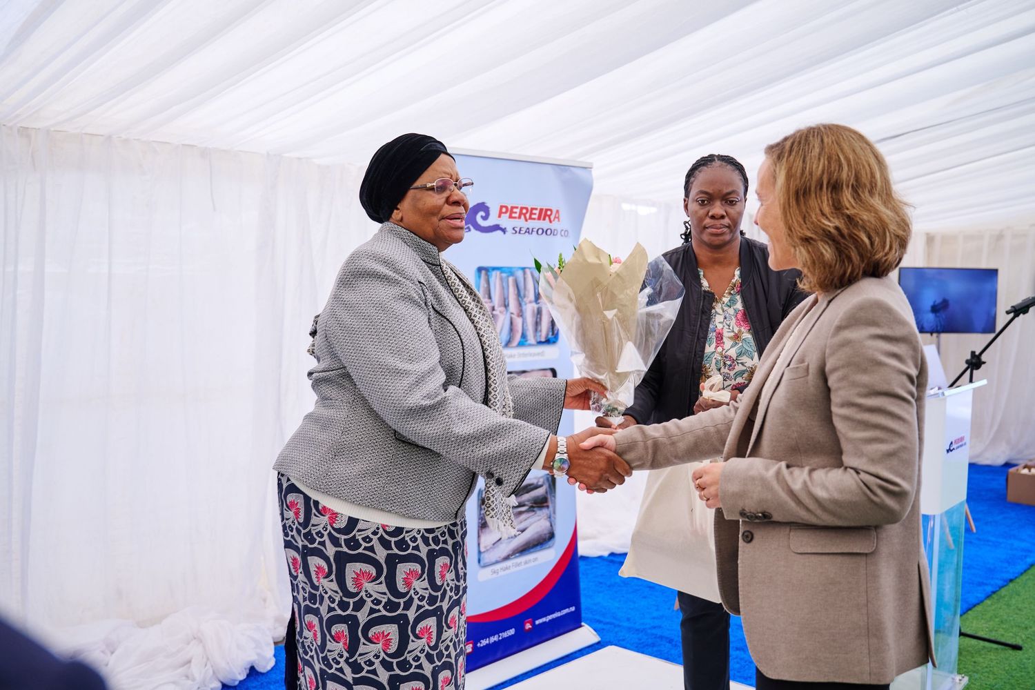 Two women shaking hands at a formal event under a white tent, with a third woman standing nearby.