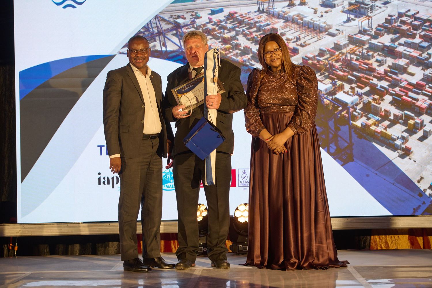 Three professionals pose together at a port industry awards ceremony with a shipping container terminal backdrop.