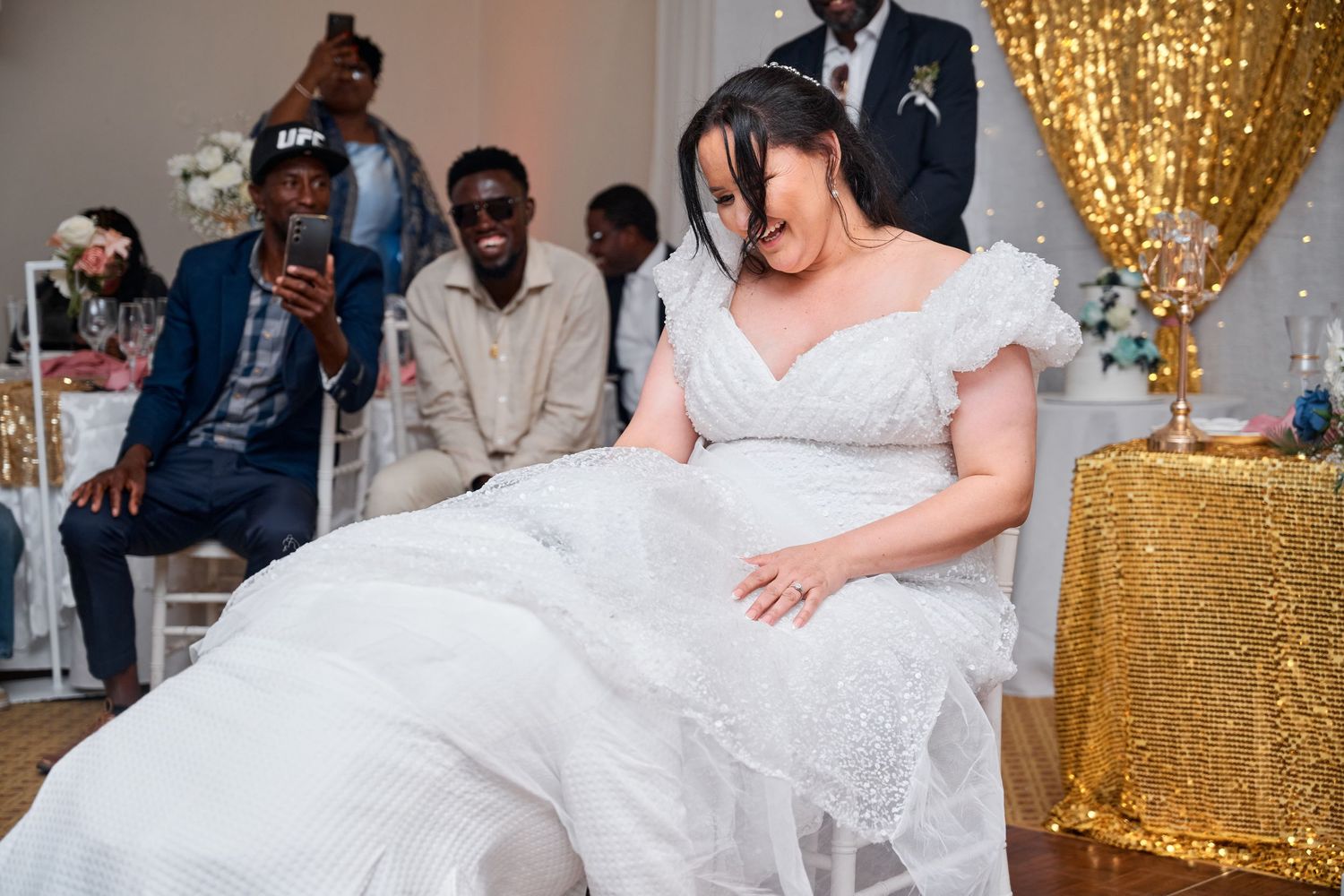 Bride in white wedding gown dancing joyfully at reception with guests celebrating in gold-decorated venue.