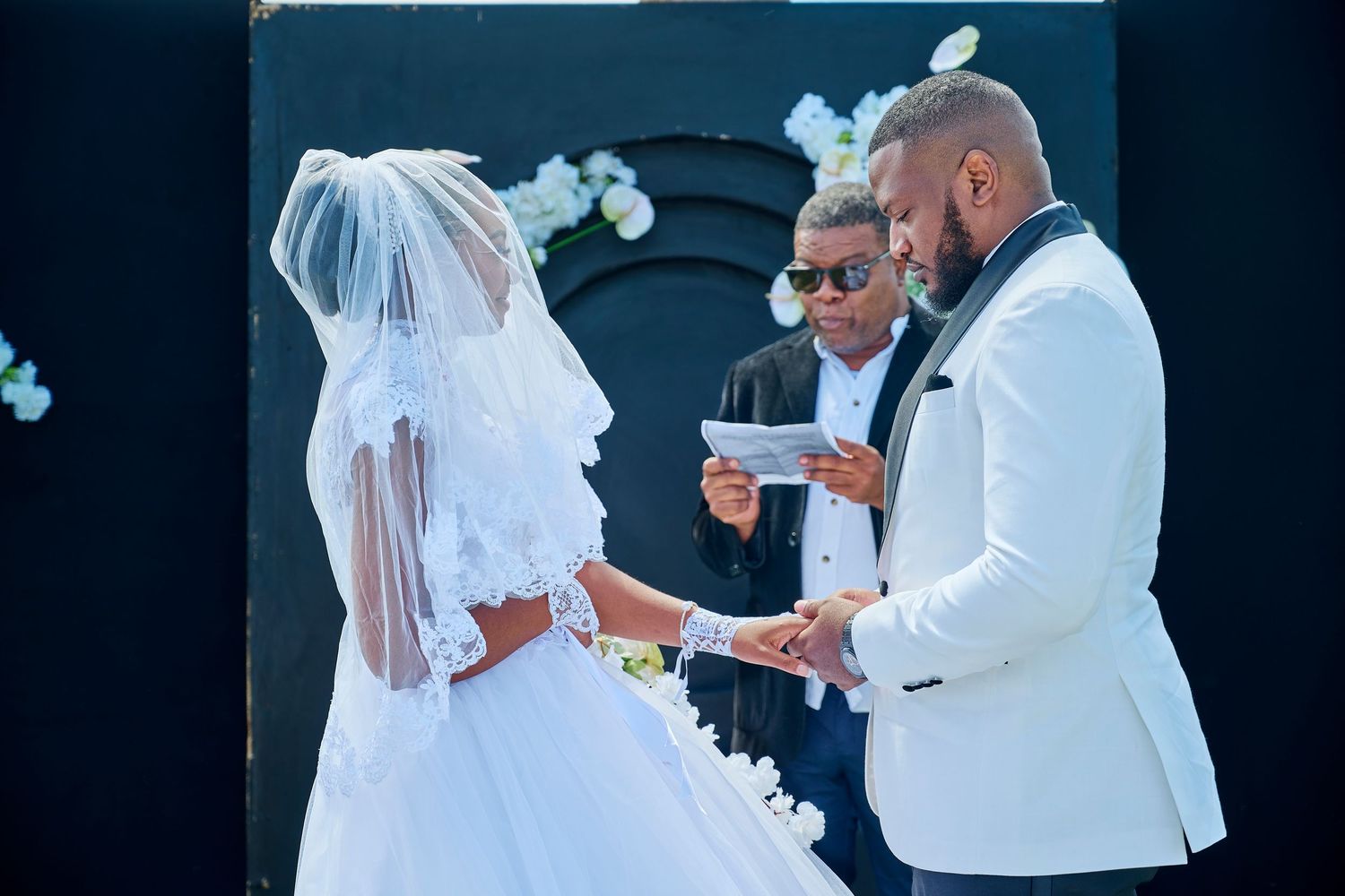 A groom in a white tuxedo exchanges vows with his bride in a white wedding gown and veil at an outdoor ceremony.