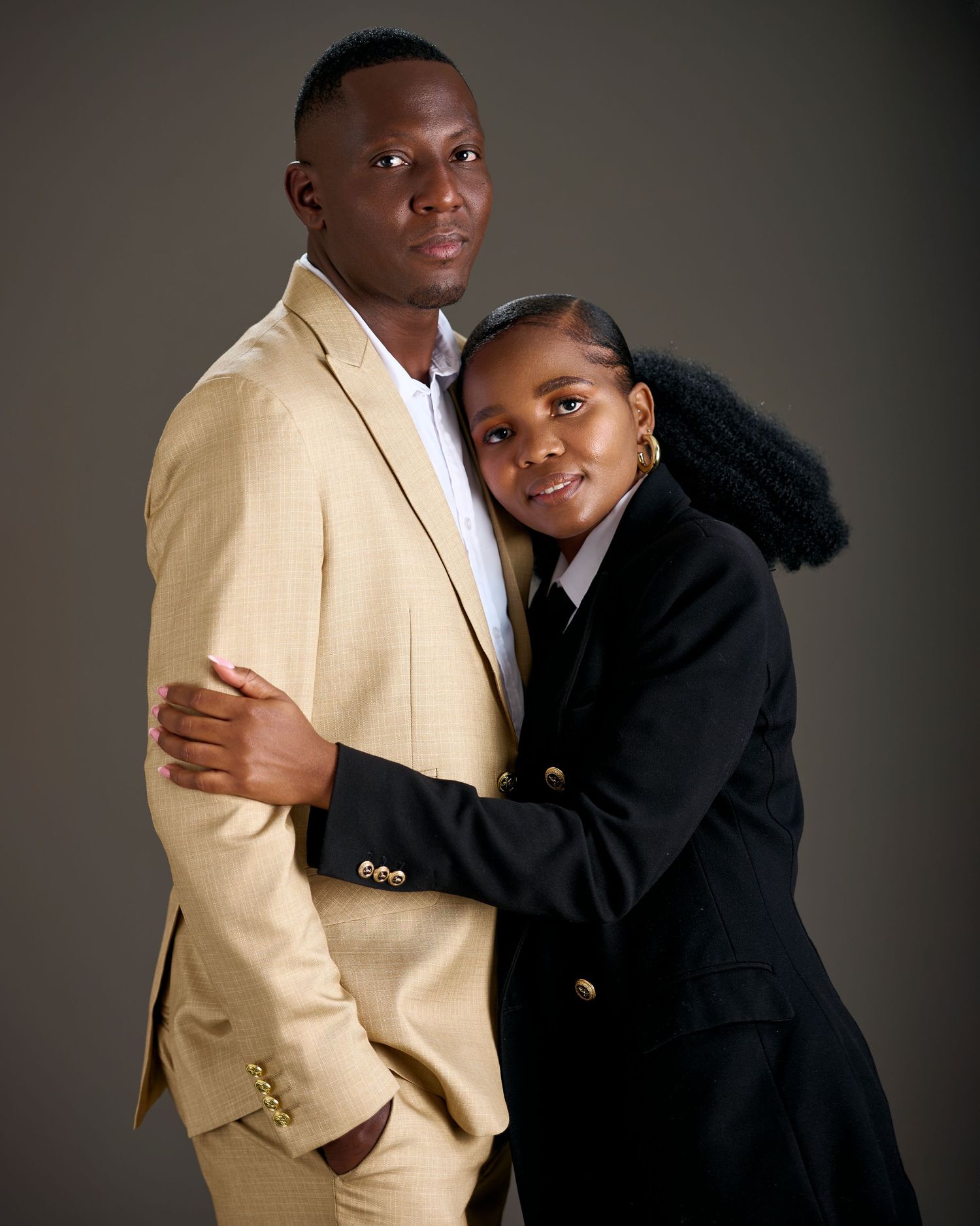 A man in a beige suit and woman in a black blazer pose together in a professional studio portrait.