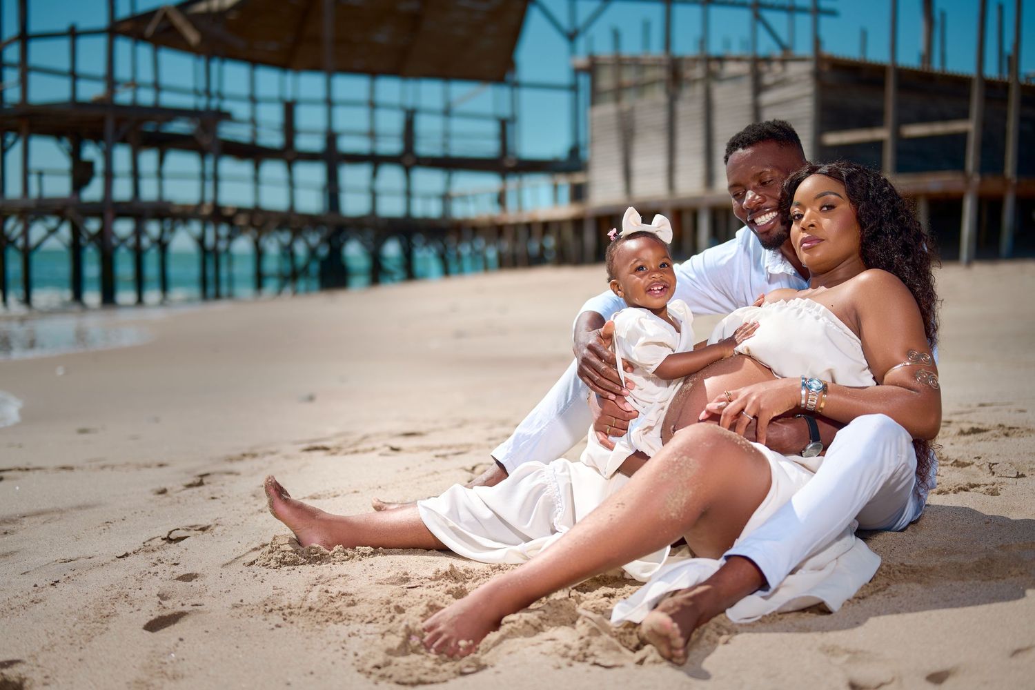 Family of three sitting on sandy beach, parents in white outfits holding baby girl, wooden pier in background.