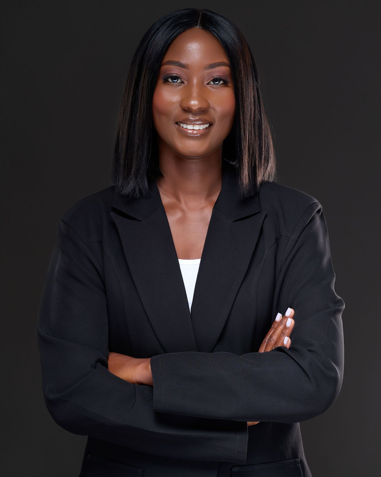 Professional woman in black blazer smiling with arms crossed against dark background.
