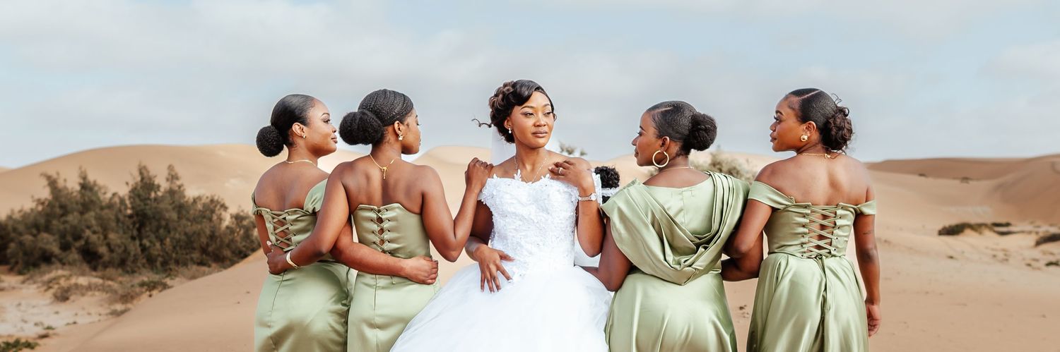 Bride in white gown poses with bridesmaids in sage green dresses against sandy desert dunes backdrop.