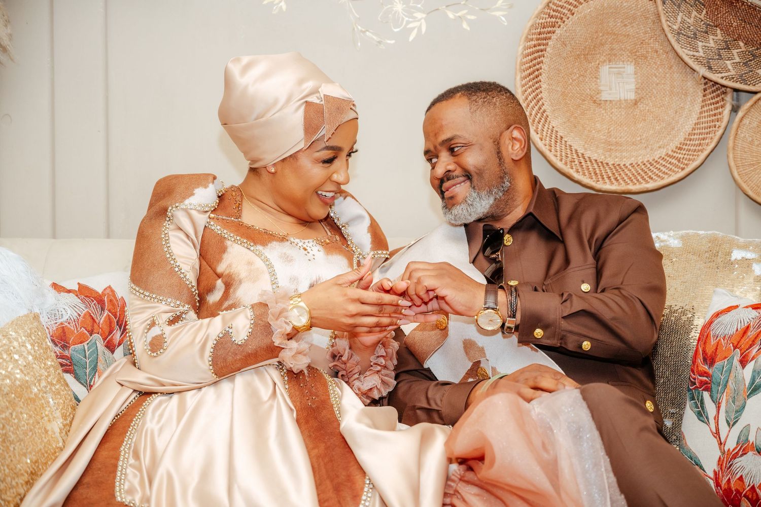 Couple in traditional African attire exchanging rings during a wedding ceremony, seated on a decorated sofa.