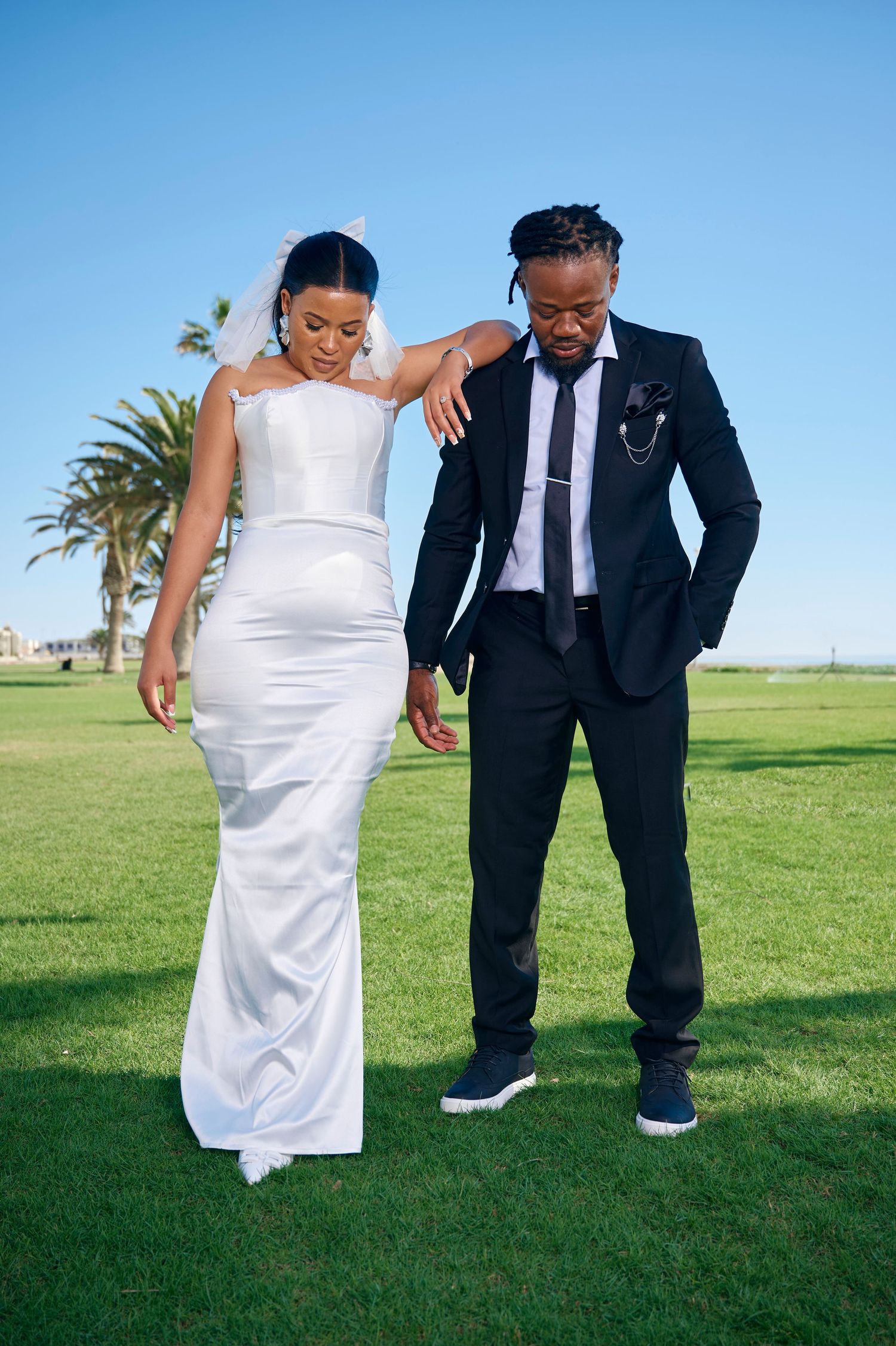 A bride in a white gown and groom in a black suit stand on green grass under a clear blue sky.