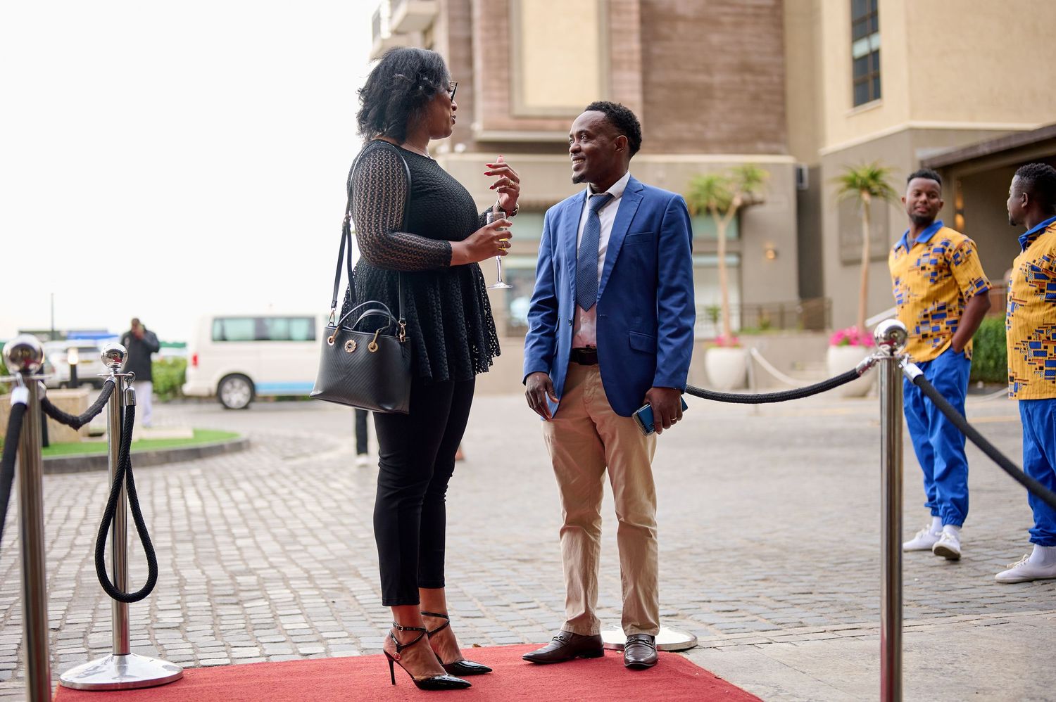 Two people in formal attire converse on a red carpet outside a modern building at a professional event.