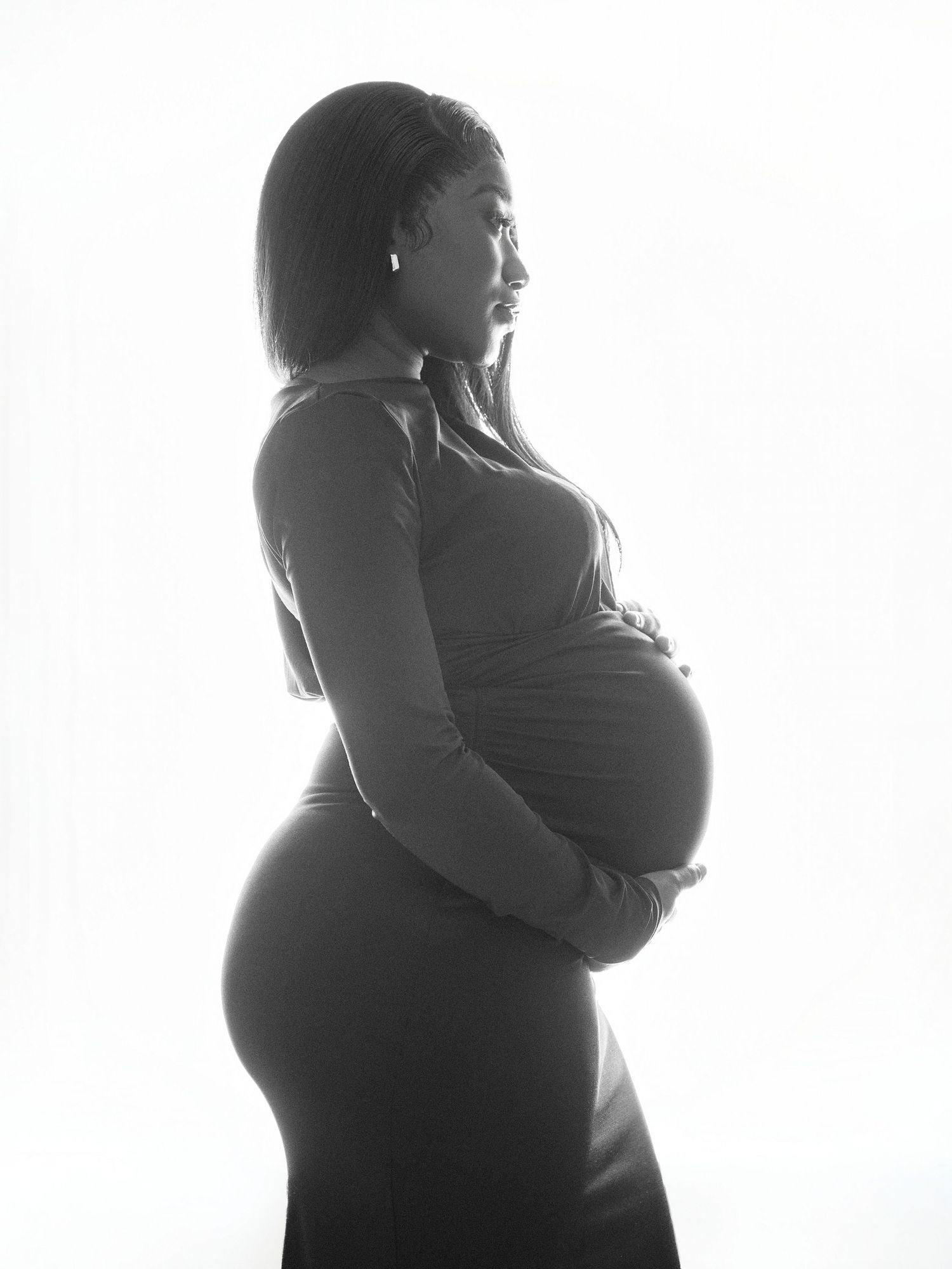 Black and white maternity photo of a pregnant woman in profile, cradling her baby bump in a fitted long-sleeve dress.