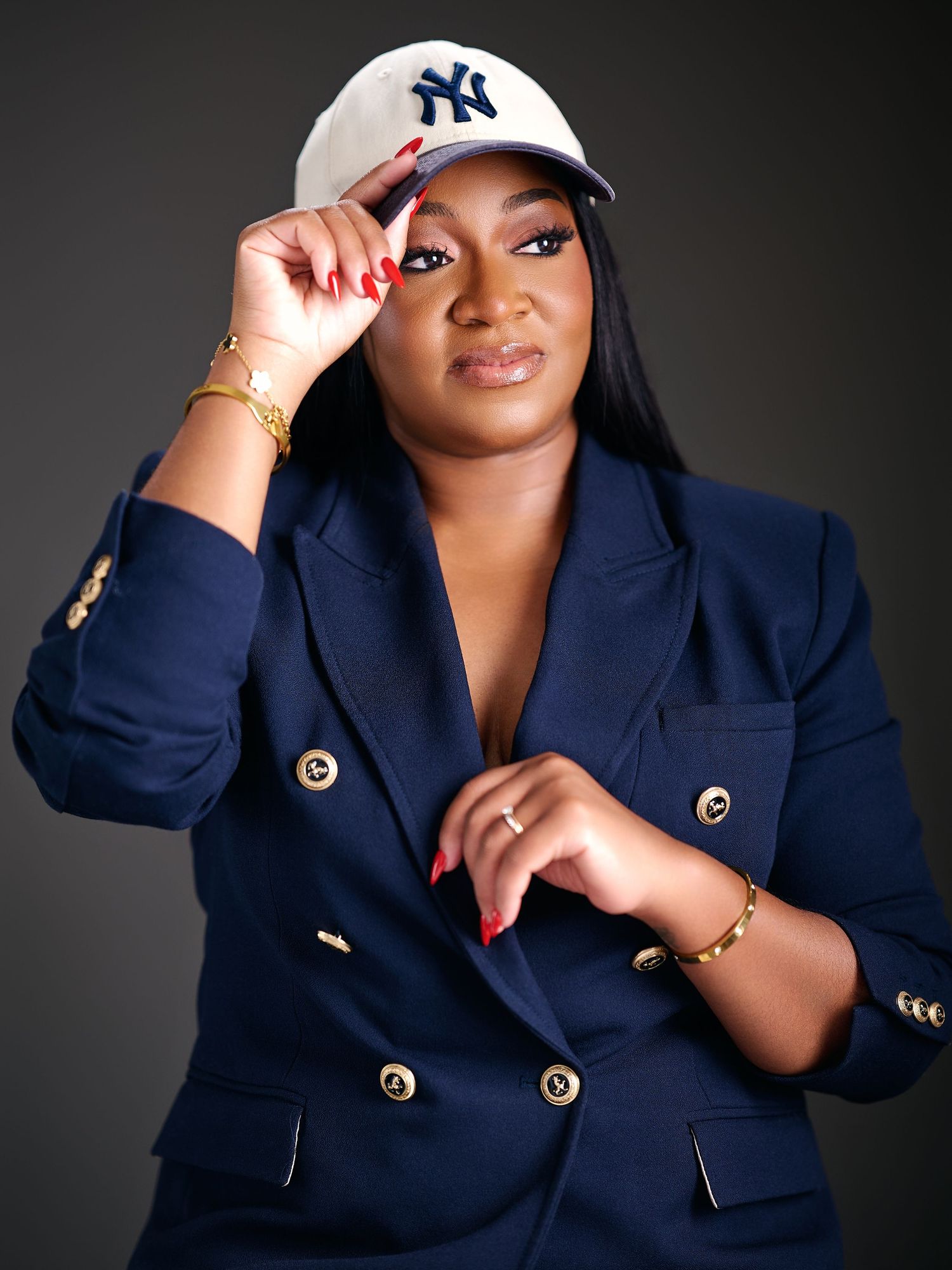 Woman in navy blazer and NY Yankees cap poses confidently, wearing gold jewelry with red nails.