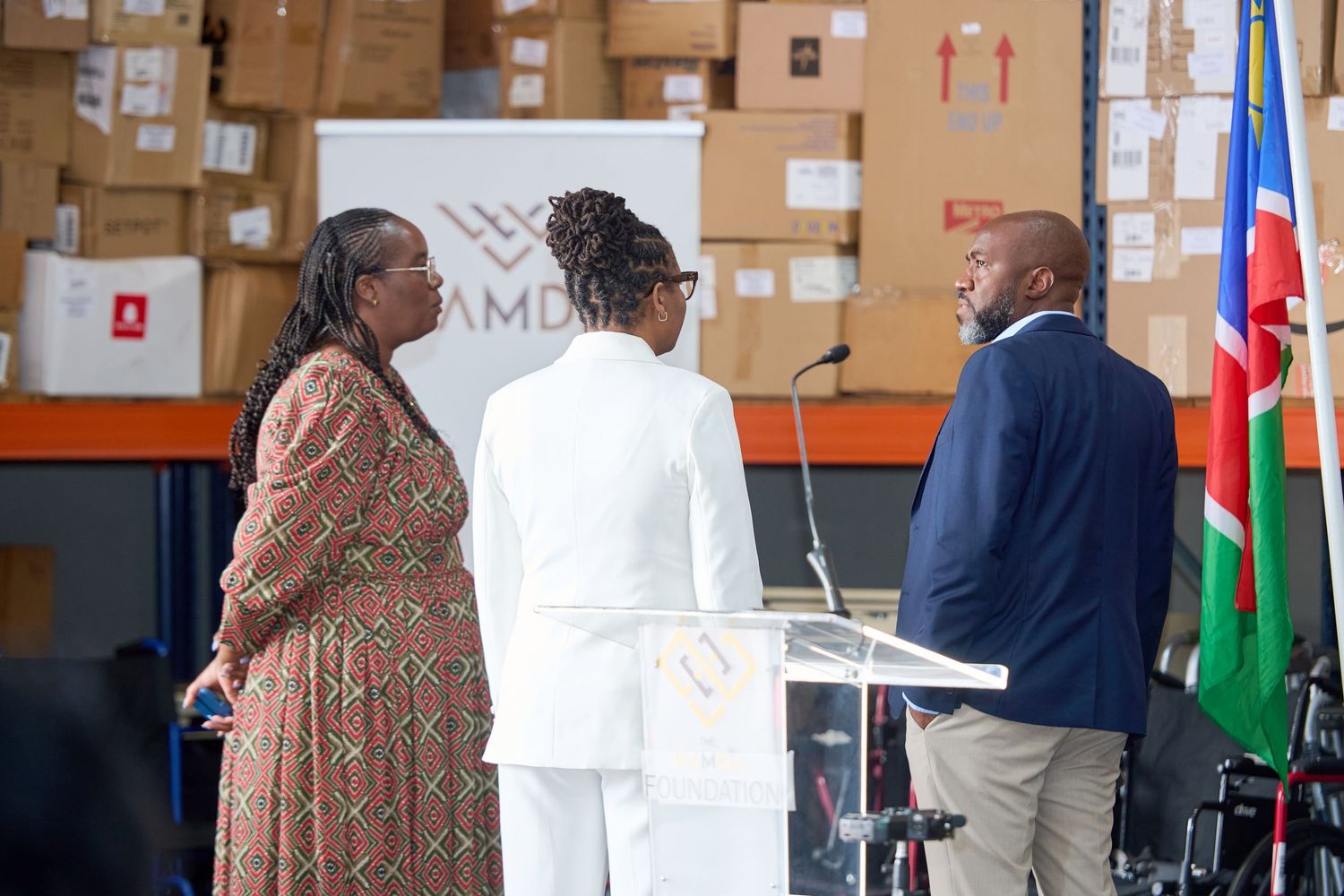 Three professionals conversing near a podium in a warehouse with boxes and a Namibian flag in the background.