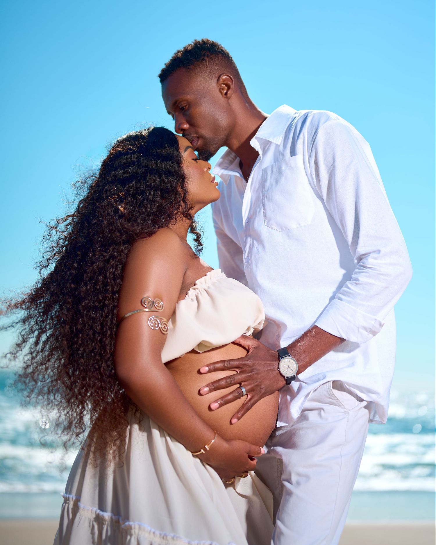 Couple sharing a tender moment on the beach, woman in white maternity dress with curly hair, man in white outfit.