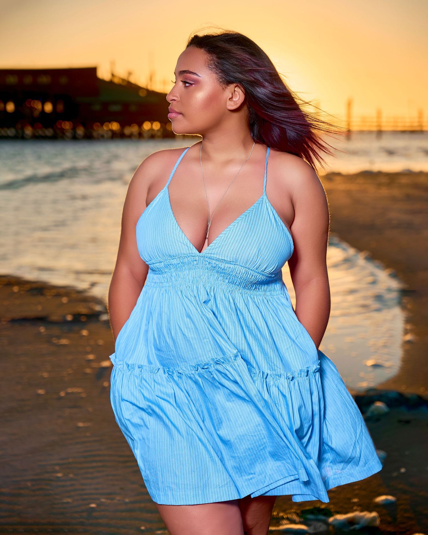Woman in flowing light blue dress poses on beach during golden sunset by the ocean.