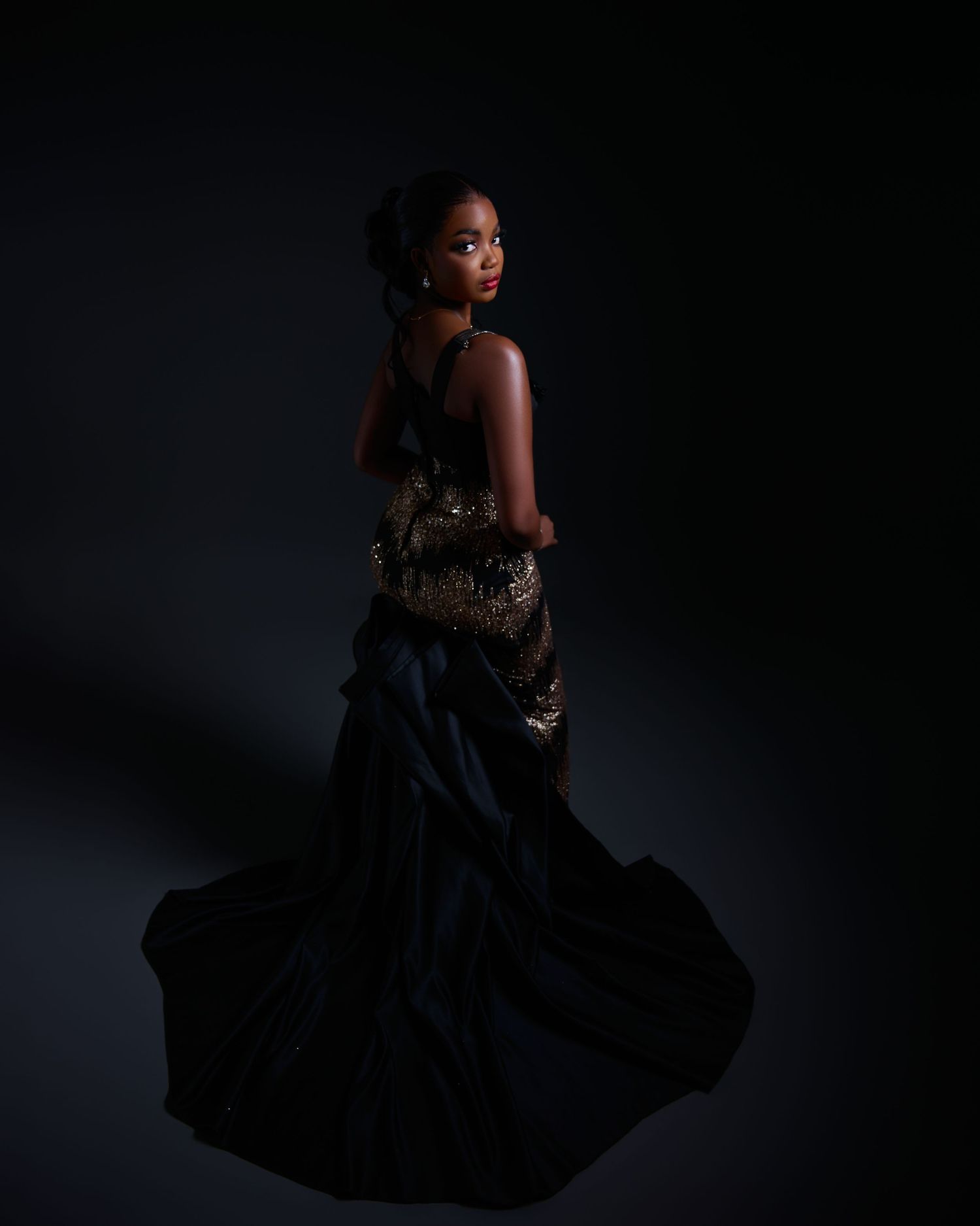 Woman in elegant black and gold gown poses gracefully against dark studio background.