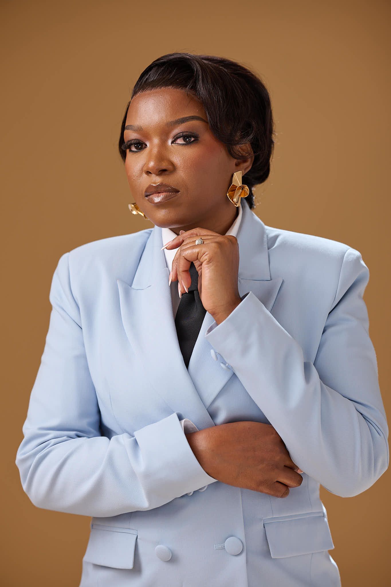 A confident Black woman in a light blue blazer poses thoughtfully with gold earrings against a warm brown background.