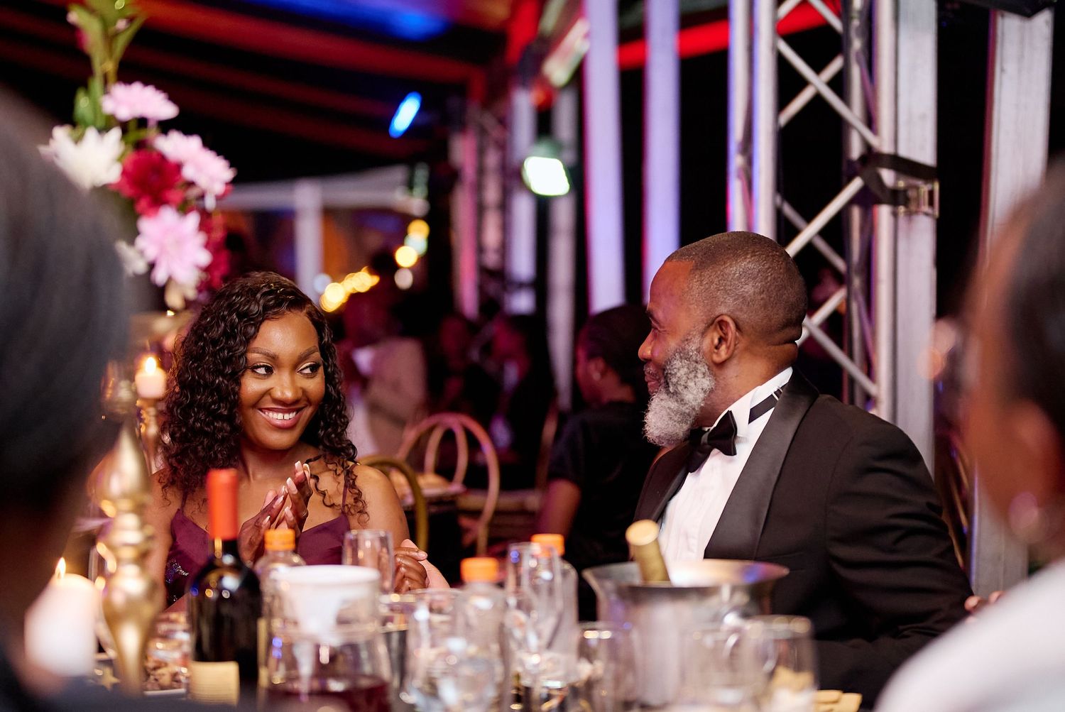 Elegant couple sharing a conversation at a formal dinner event, woman in burgundy dress smiling at man in black tuxedo.