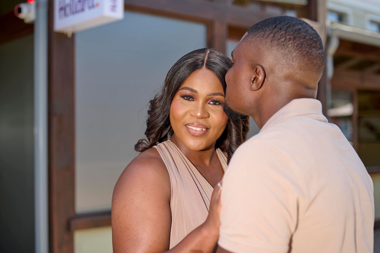 A couple sharing a romantic moment, man kissing woman's cheek outdoors, both dressed in elegant beige attire.