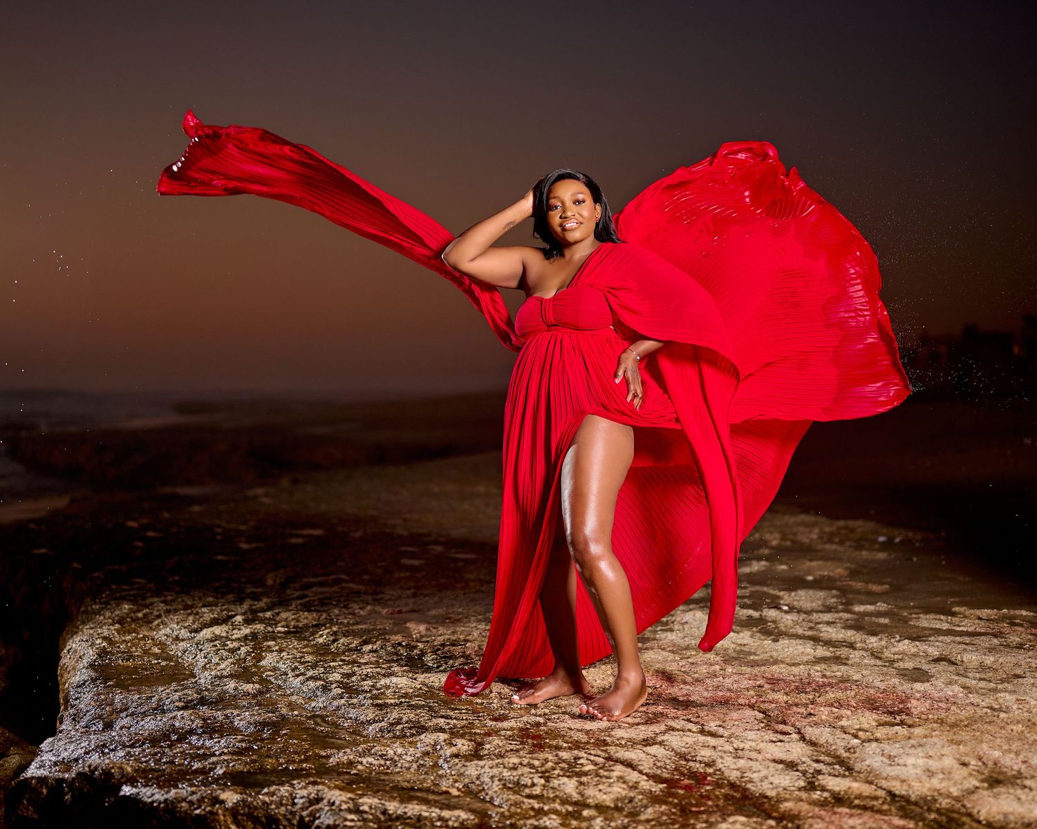 Woman in flowing red gown poses dramatically on rocks at night with fabric billowing in the wind.