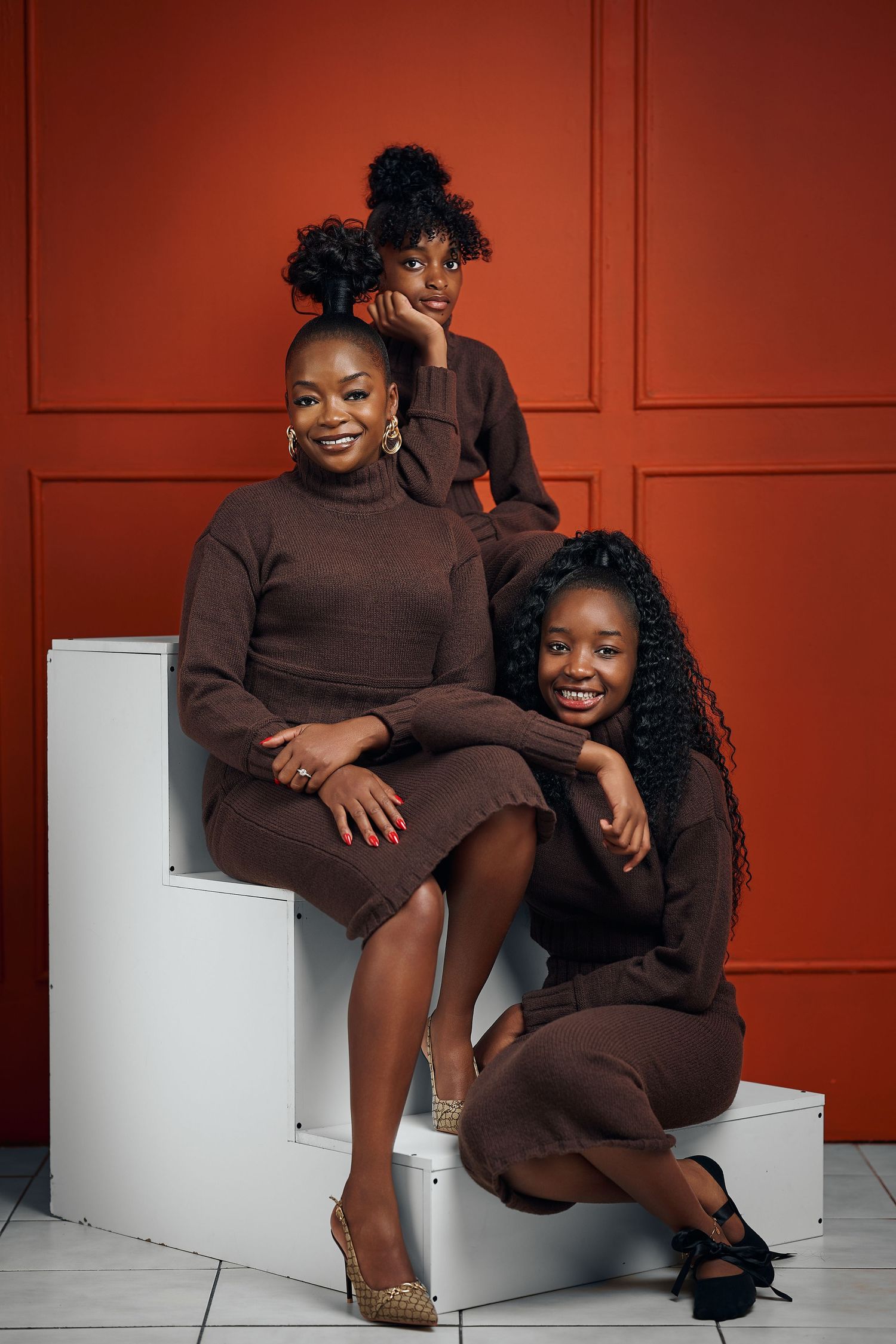 Three women in matching brown sweater dresses pose together against a red wall in a professional studio portrait.