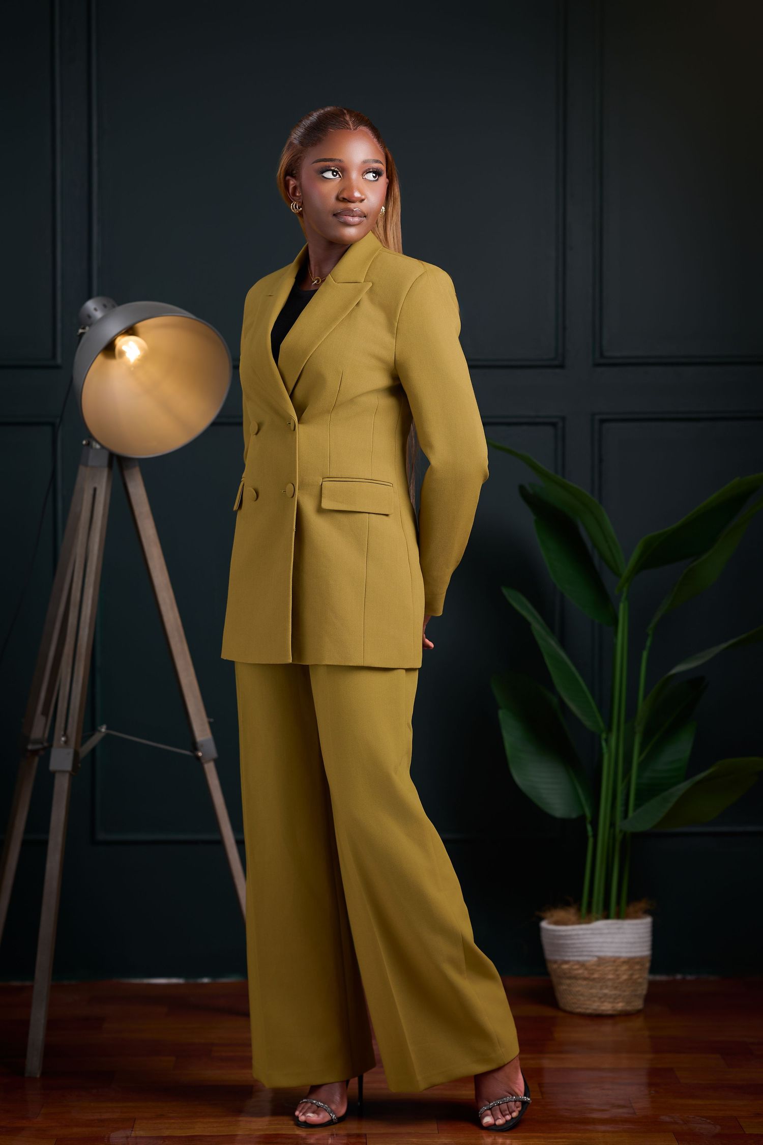 Woman in olive green pantsuit posing confidently in a dark green studio with a floor lamp and plant.