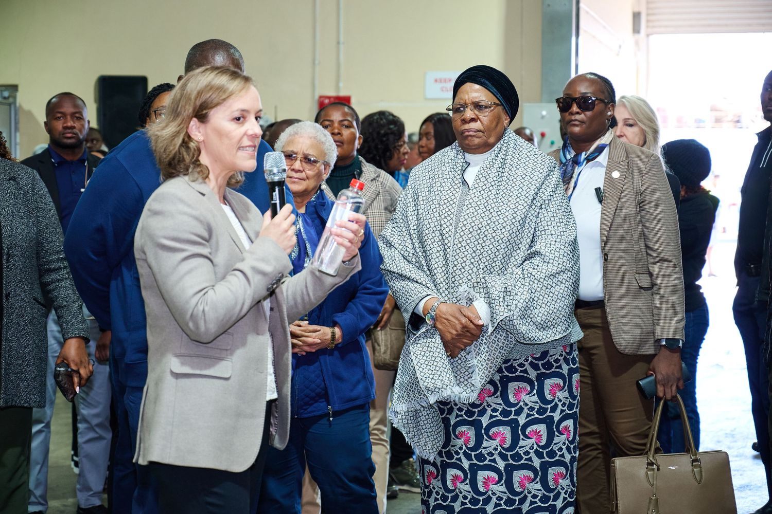 A woman in a gray blazer speaks to a group of diverse professionals gathered indoors at a formal event.