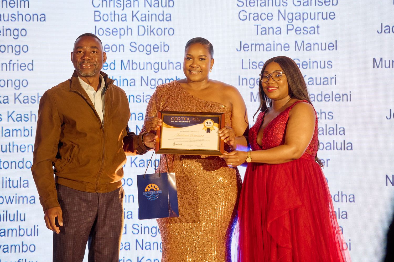 Woman in gold dress receives certificate award from two presenters at a formal recognition ceremony.