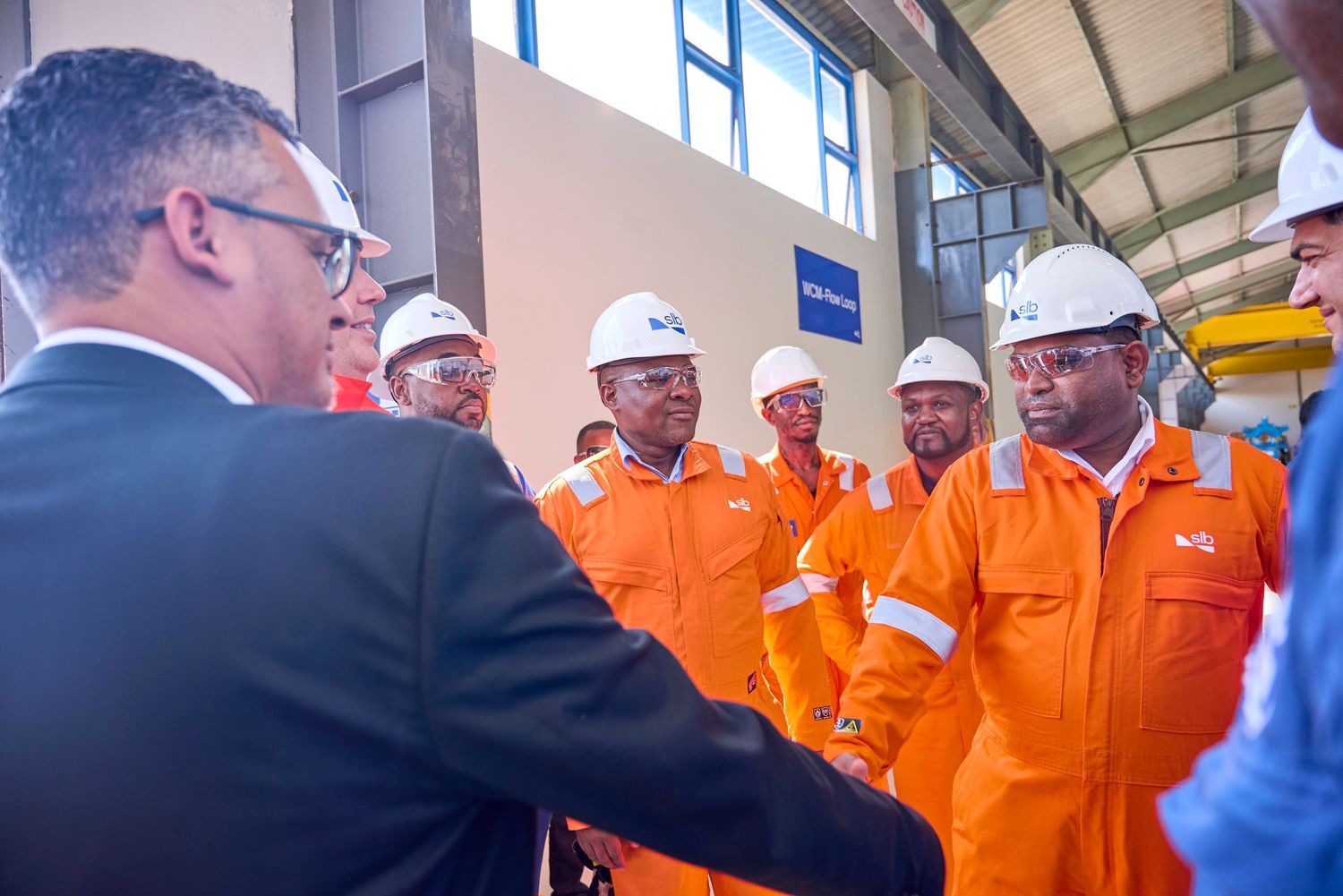 Workers in orange safety gear and white helmets receive a briefing from a suited official at an industrial facility.