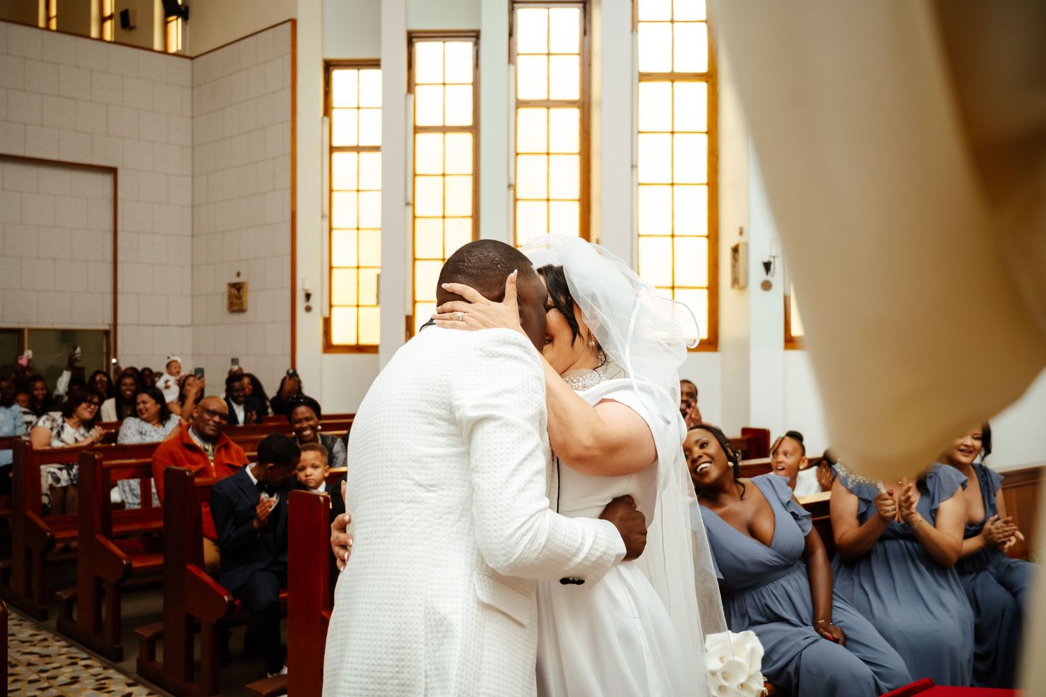 Bride and groom share first kiss at church altar during wedding ceremony with guests applauding in pews.