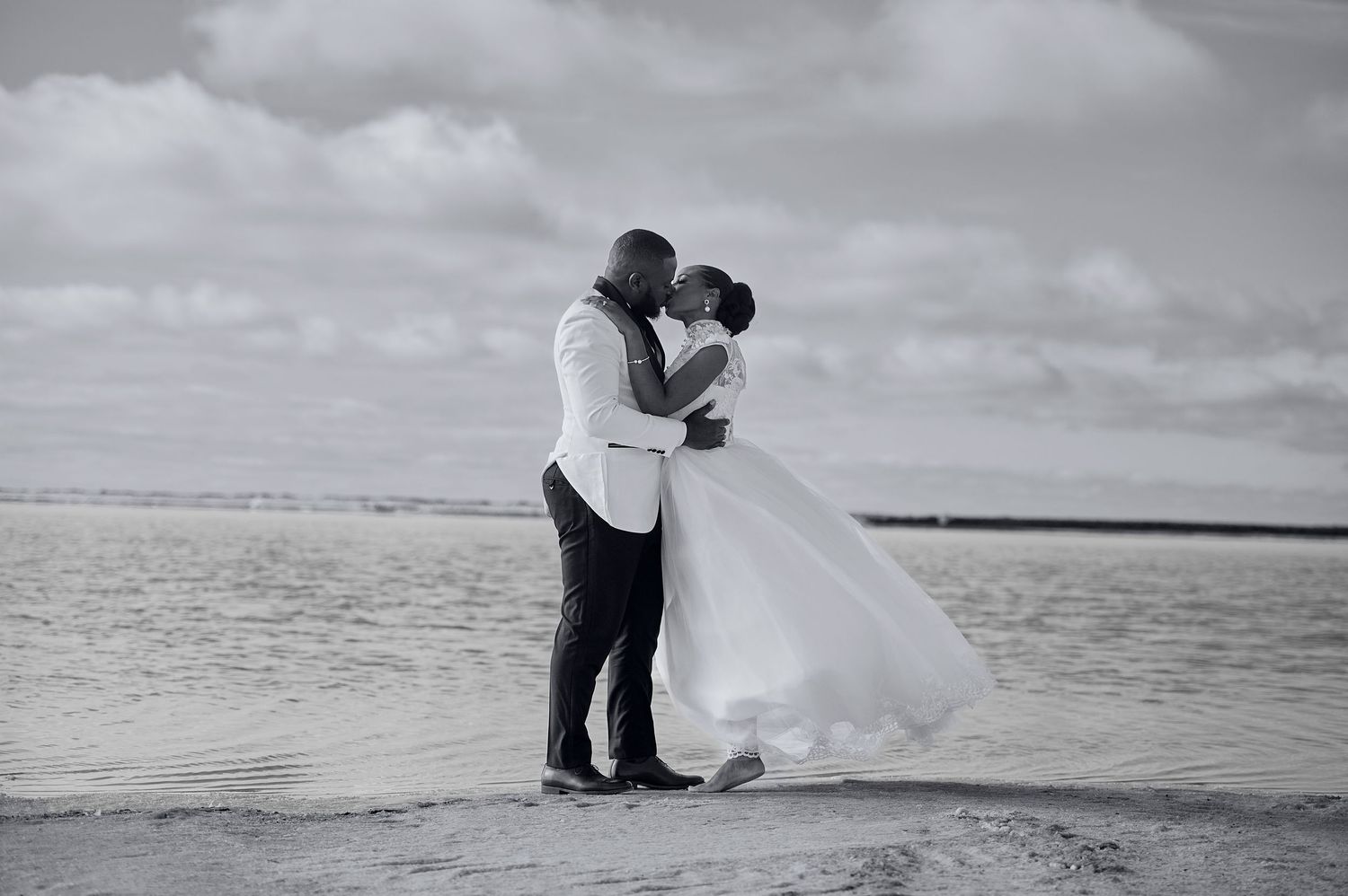 Bride and groom share a romantic kiss on the beach in a stunning black and white wedding photo.