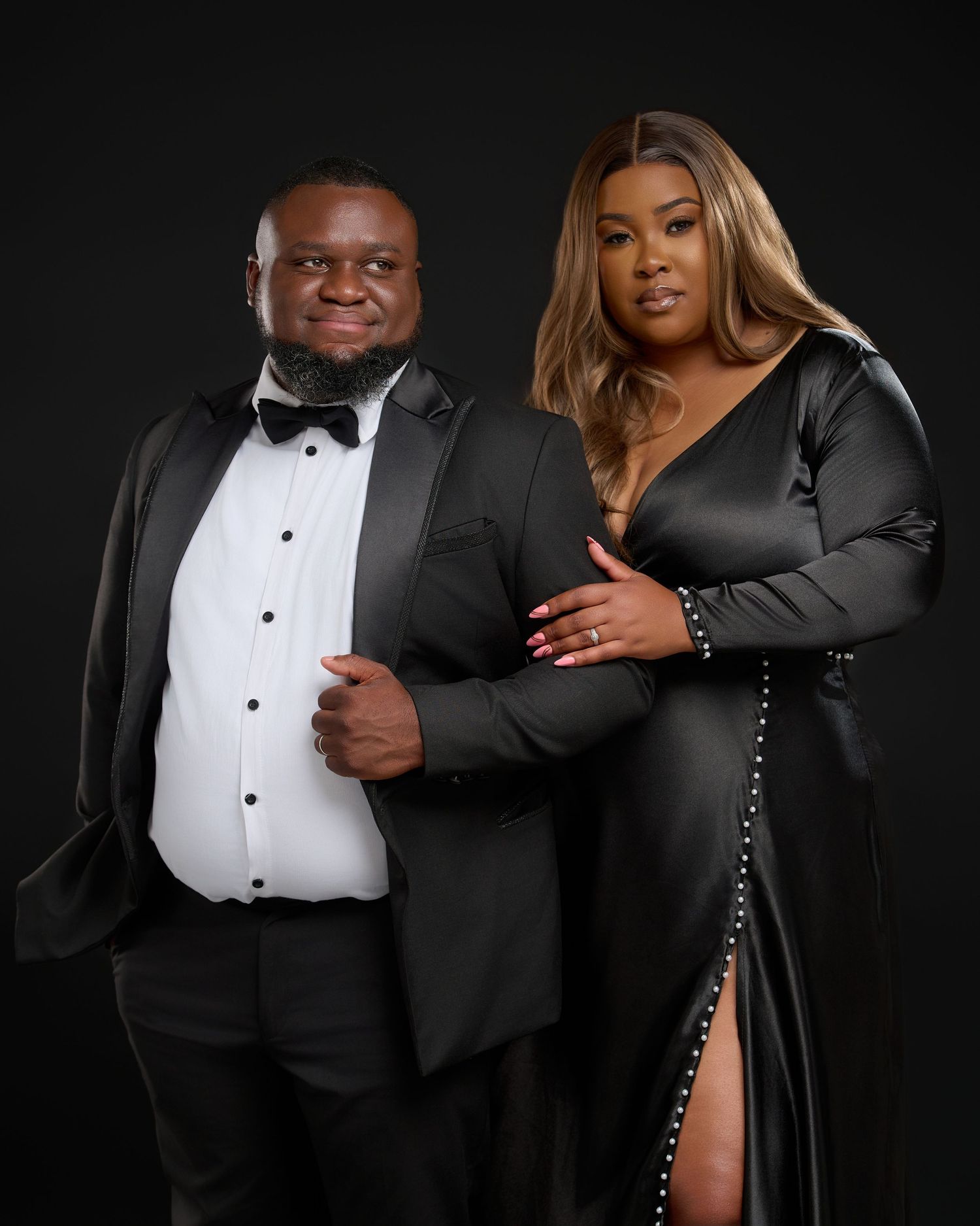 Elegant couple posing in matching black formal attire, man in tuxedo and woman in sleek gown, against dark studio backdrop.