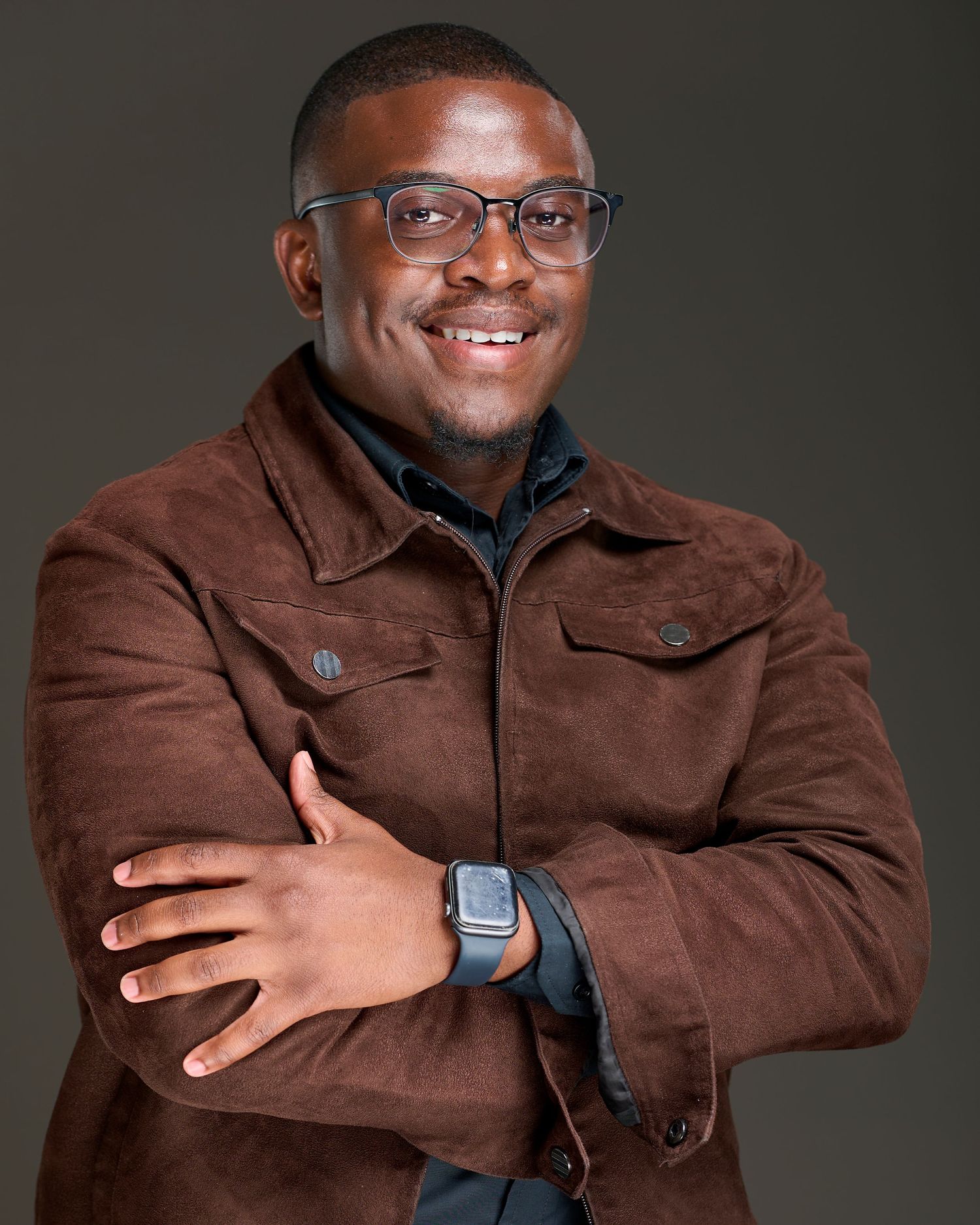 Person wearing a brown suede jacket and glasses poses with crossed arms against a dark background.