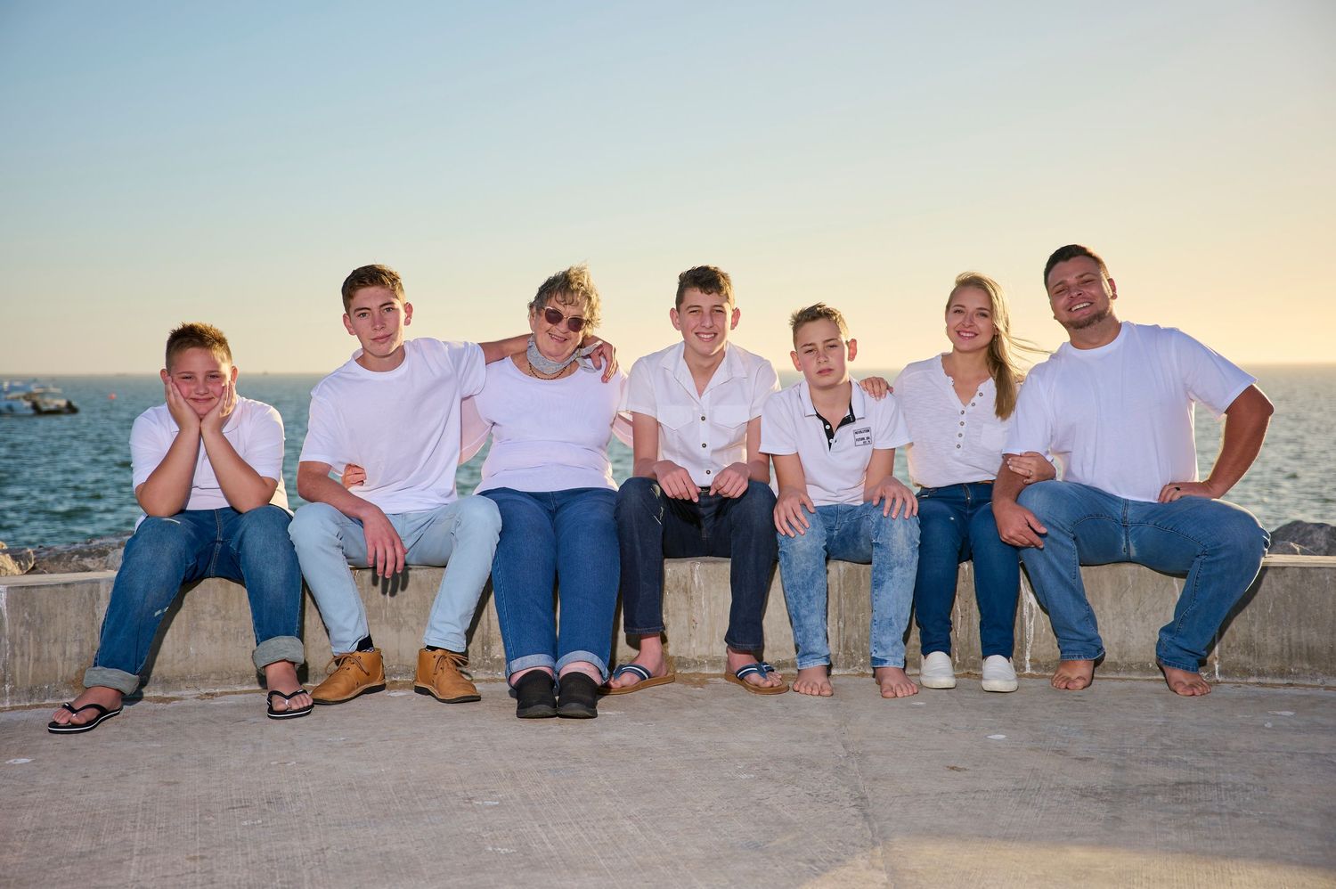 A group in casual white shirts and jeans sits together on a concrete wall by the ocean at sunset.