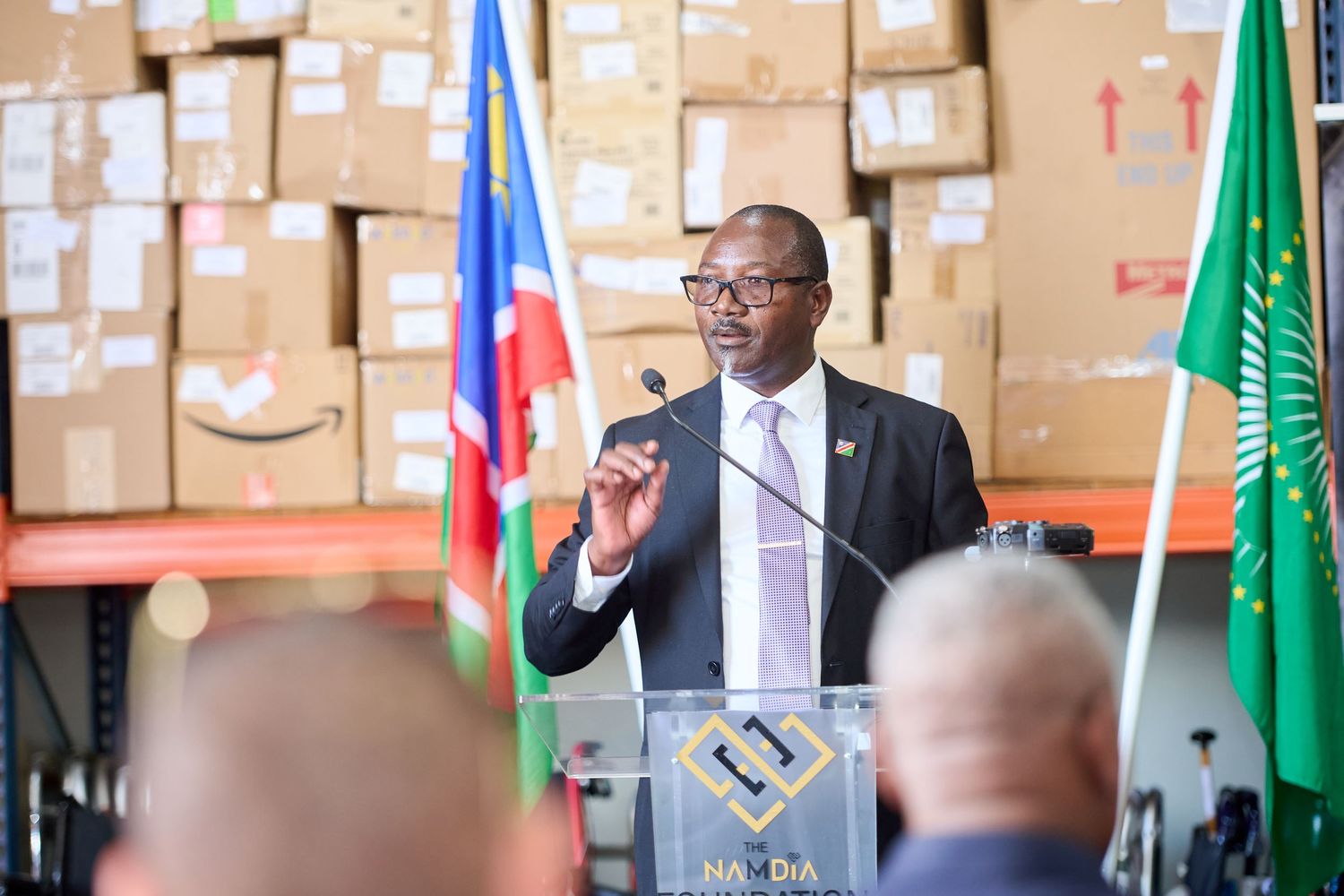 A man in a suit speaks at a podium in a warehouse with stacked boxes and African Union flag behind him.