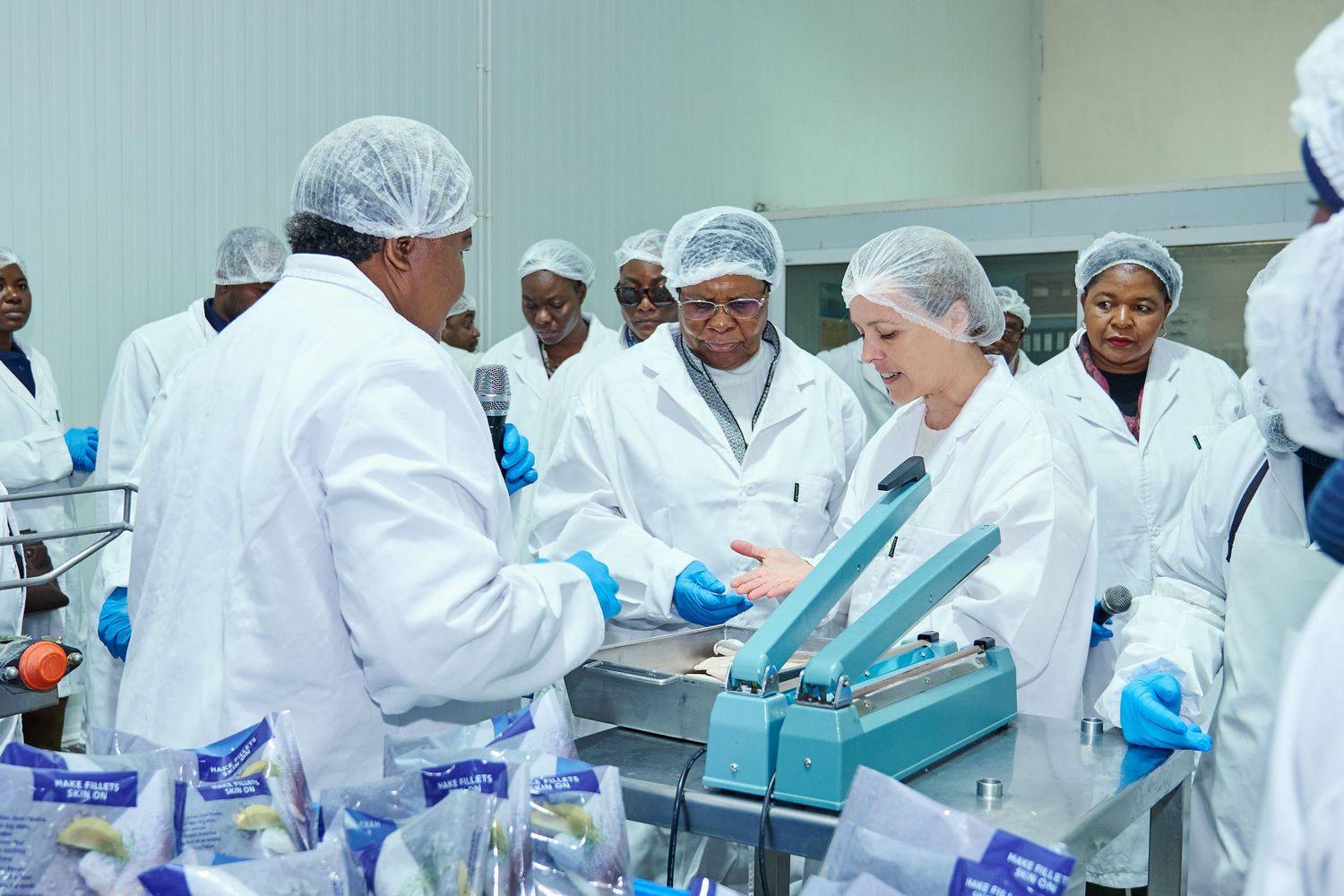 Workers in white lab coats and hairnets operating food packaging equipment in a clean manufacturing facility.