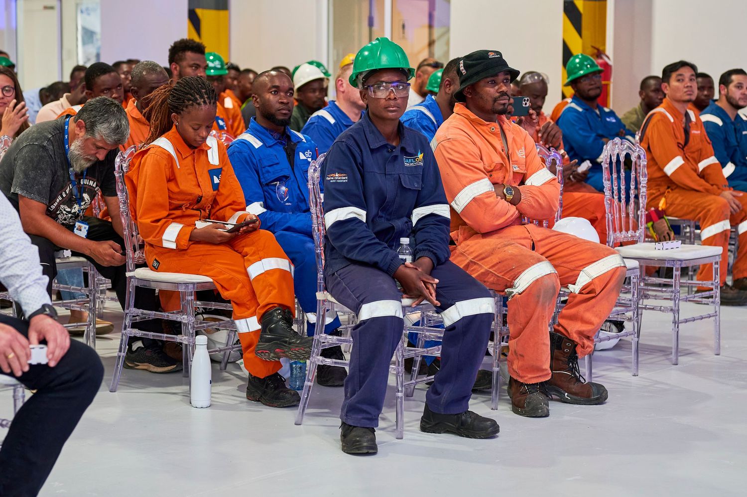Diverse workers in orange and blue coveralls attend an industrial safety briefing or company meeting.