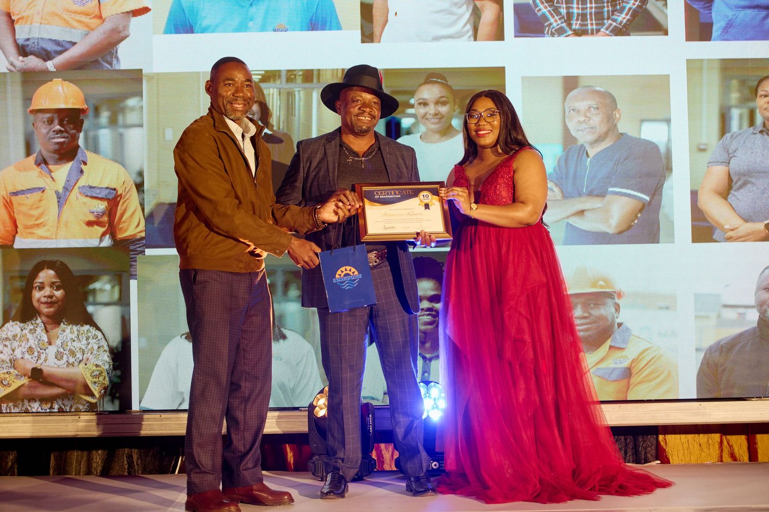 Three people on stage at an awards ceremony, with a woman in a red gown presenting a plaque to a man.
