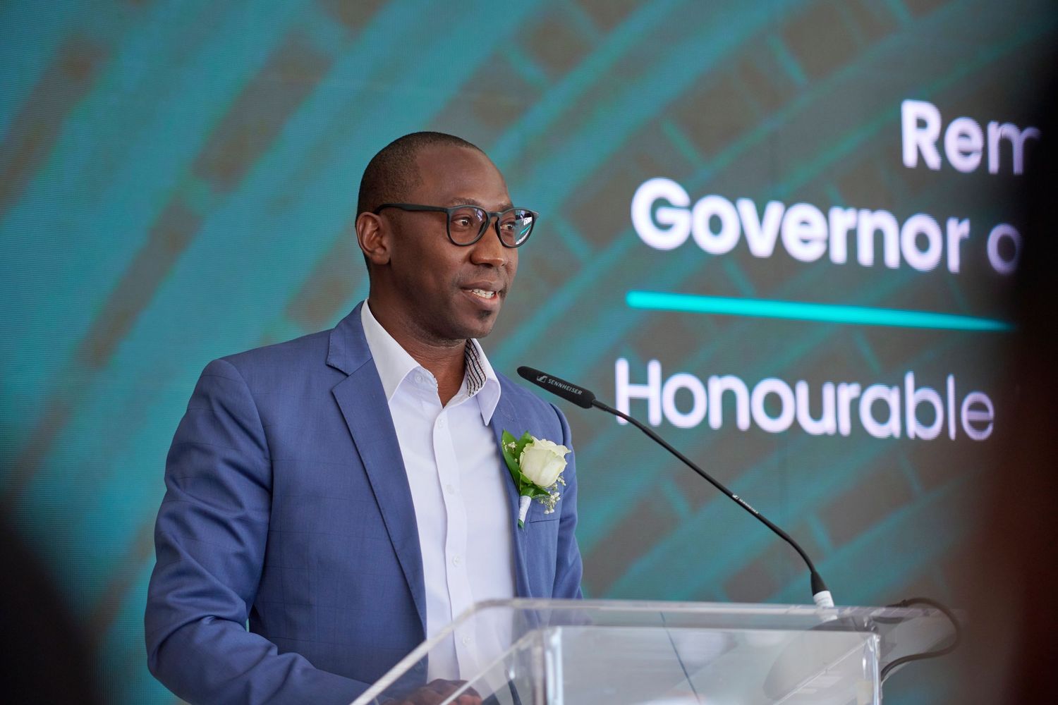 A man in a blue blazer speaks at a podium labeled Honourable Governor at a formal event.