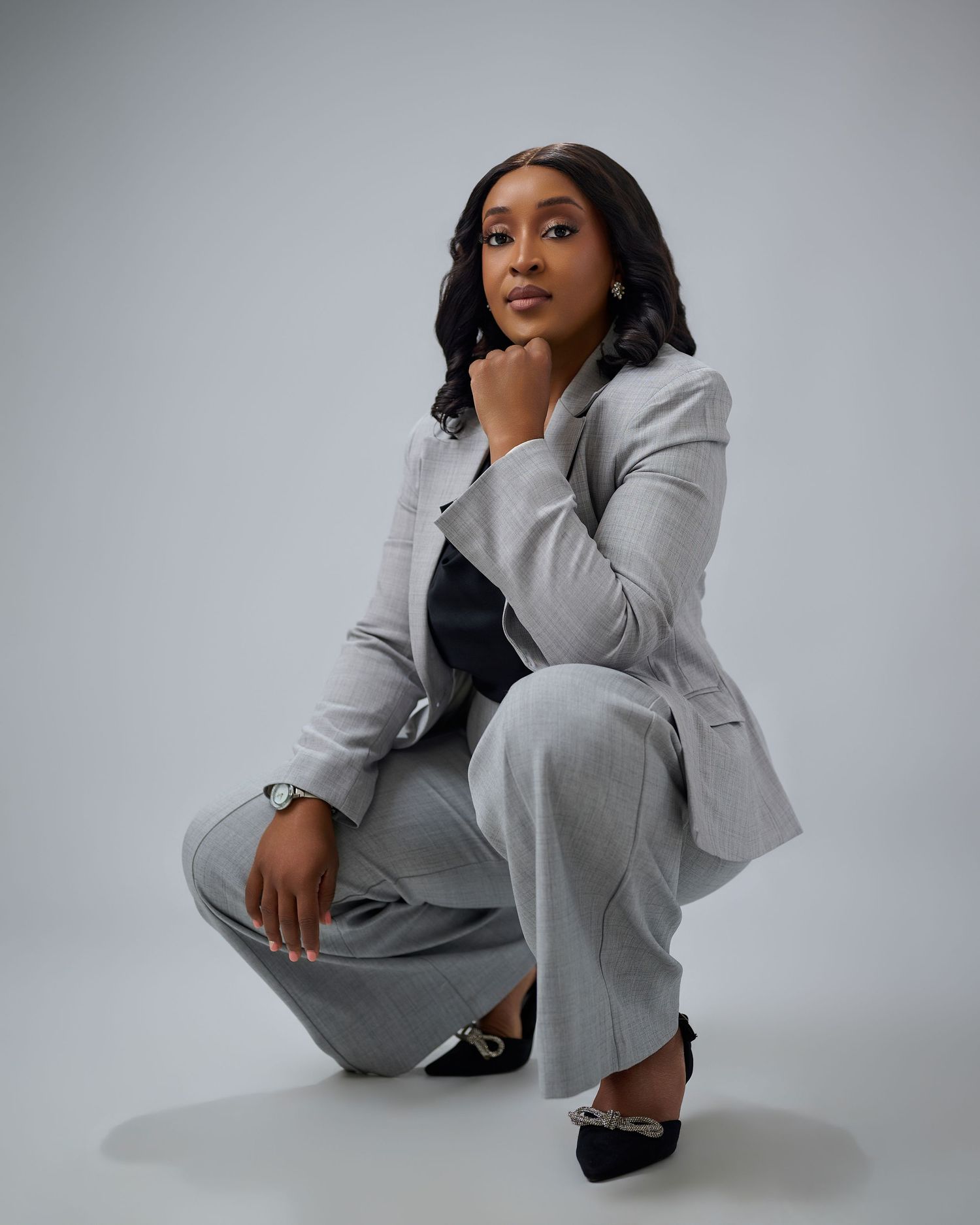 Woman in gray blazer and pants crouches confidently against white background, wearing black heels and a watch.