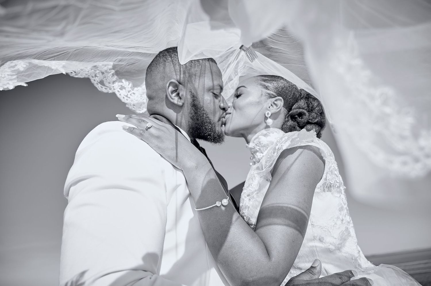 Black and white romantic photo of a bride and groom sharing a kiss beneath a flowing wedding veil.