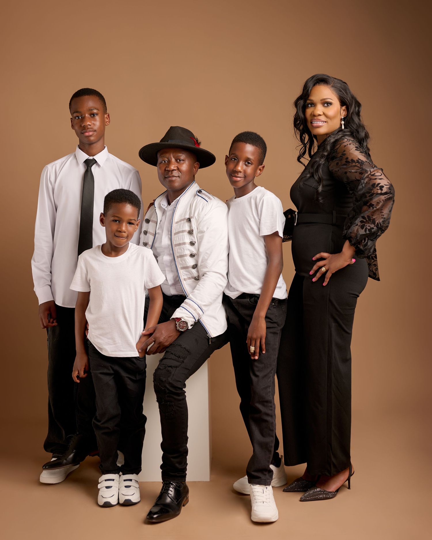 A stylish family portrait featuring parents and three sons dressed in coordinated black and white outfits against a tan background.