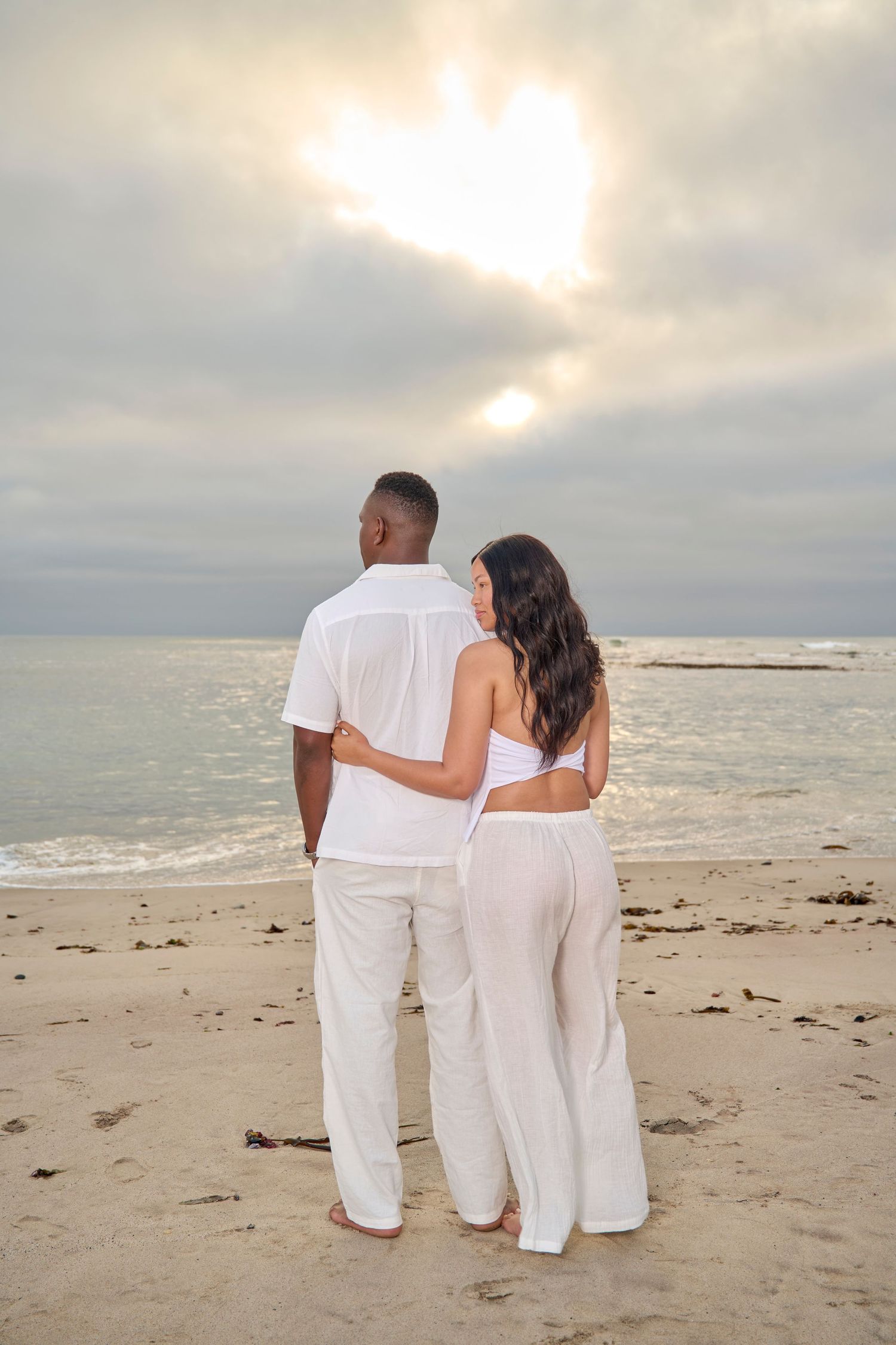 Couple in white outfits embracing on a sandy beach at sunset, gazing at the ocean under a cloudy sky.