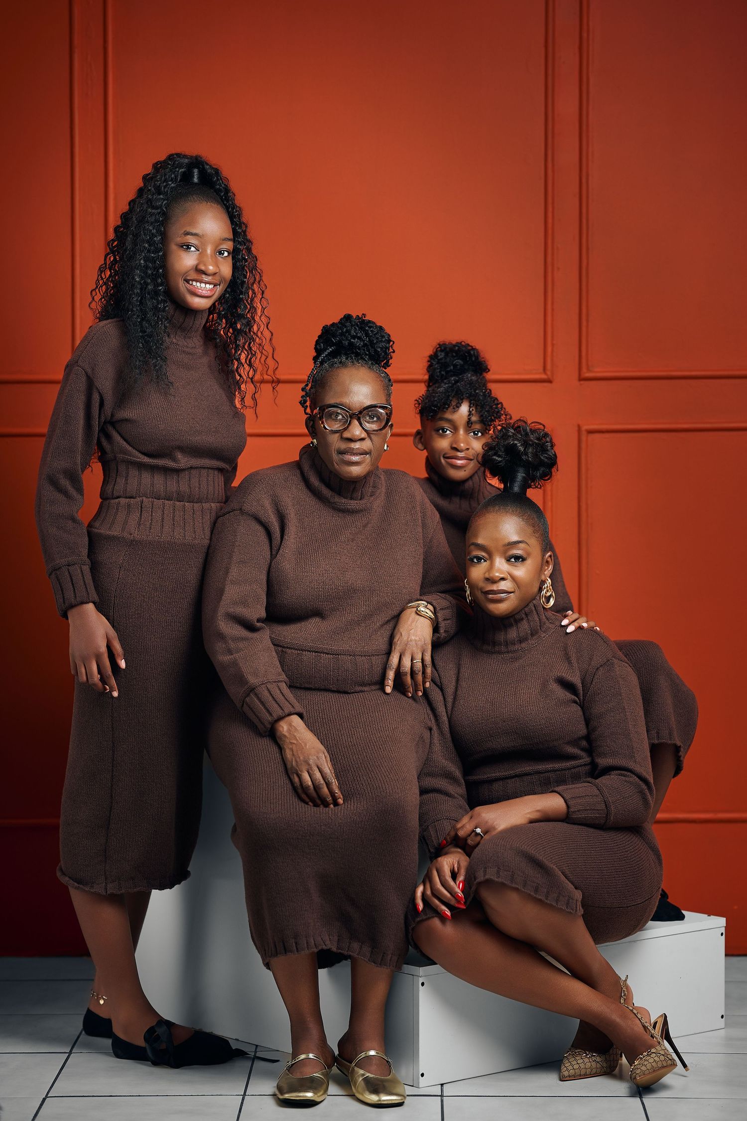 Four Black women in matching brown knit dresses pose together against a red wall in a stylish group portrait.