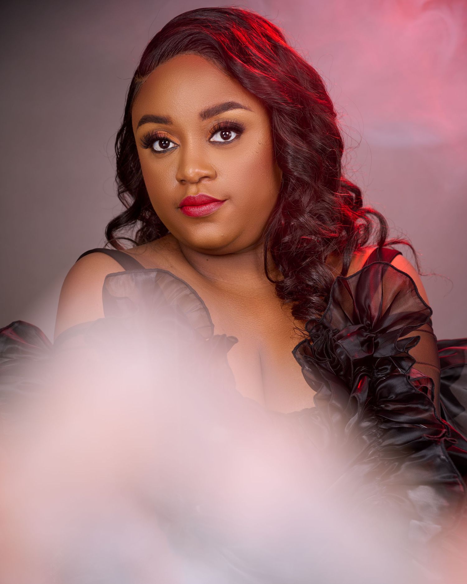 A young woman in a black ruffled off-shoulder gown poses elegantly with red lips and wavy hair in a studio.