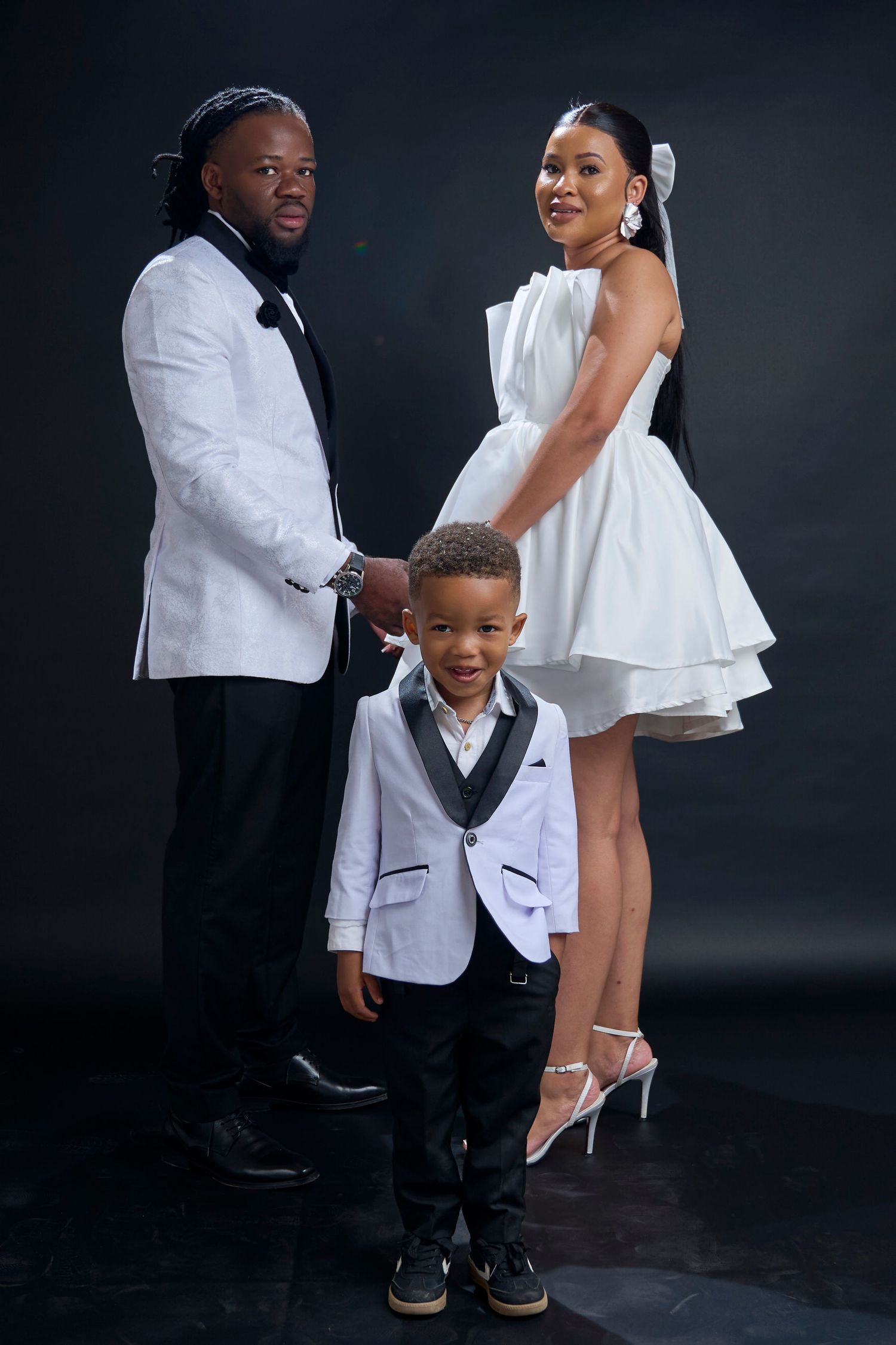 Family of three in matching white outfits posing for a formal portrait against a dark background.