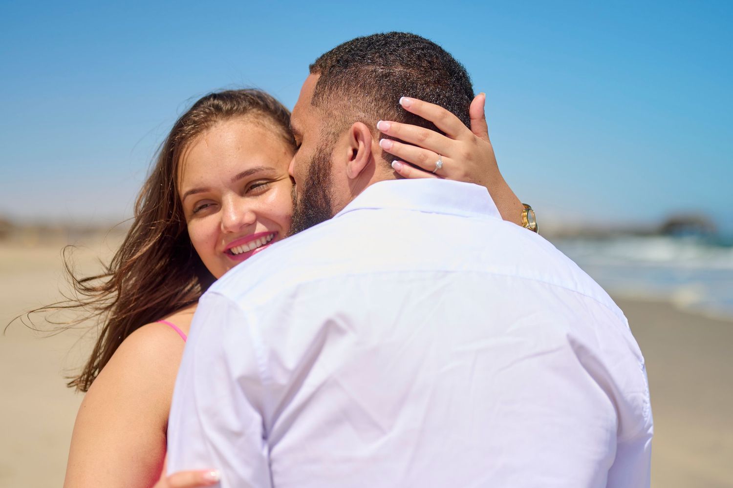 Couple embracing on a sunny beach, woman smiling over man's shoulder, ocean in the background.
