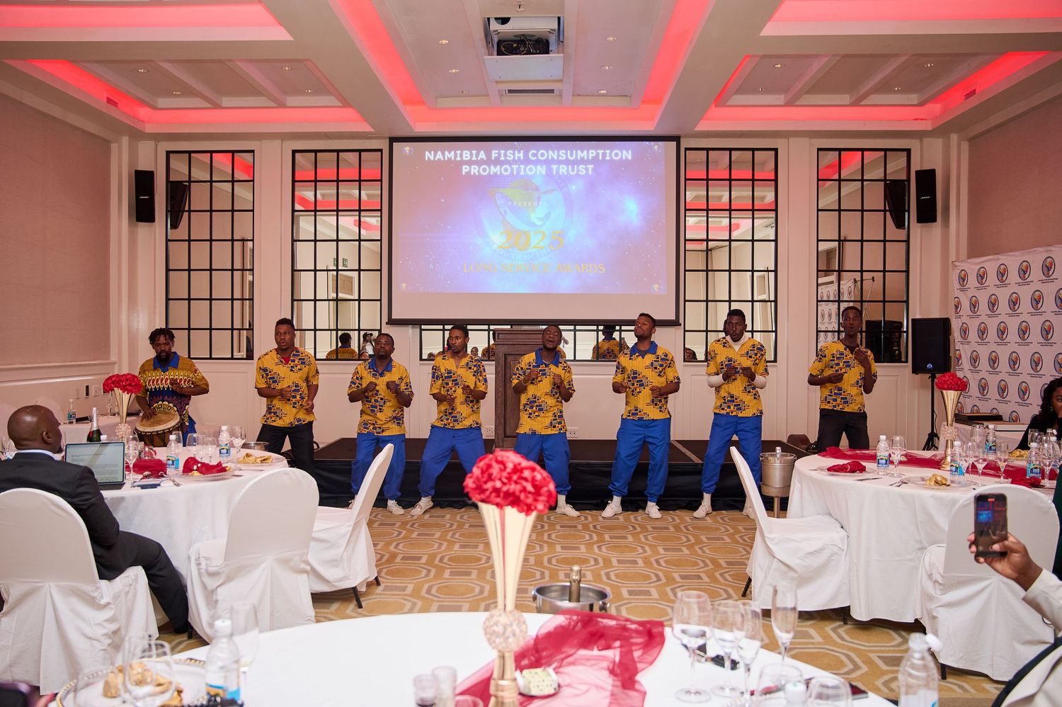 Group performing on stage at formal banquet hall with red decor and presentation screen.