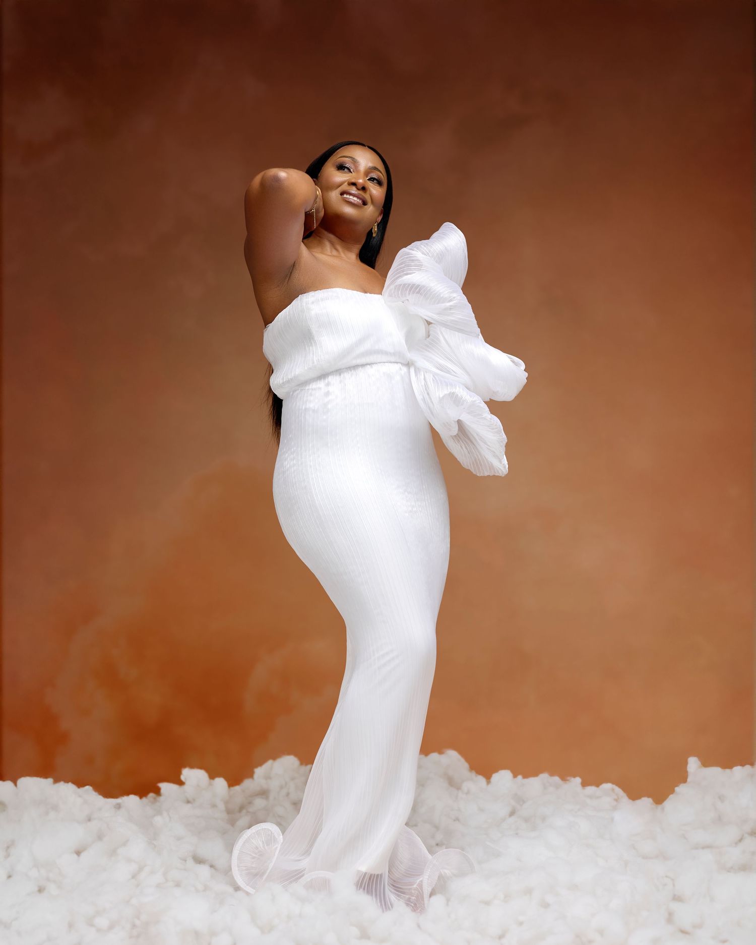 Pregnant woman in elegant white strapless gown with ruffled sleeve poses gracefully against warm brown studio backdrop.