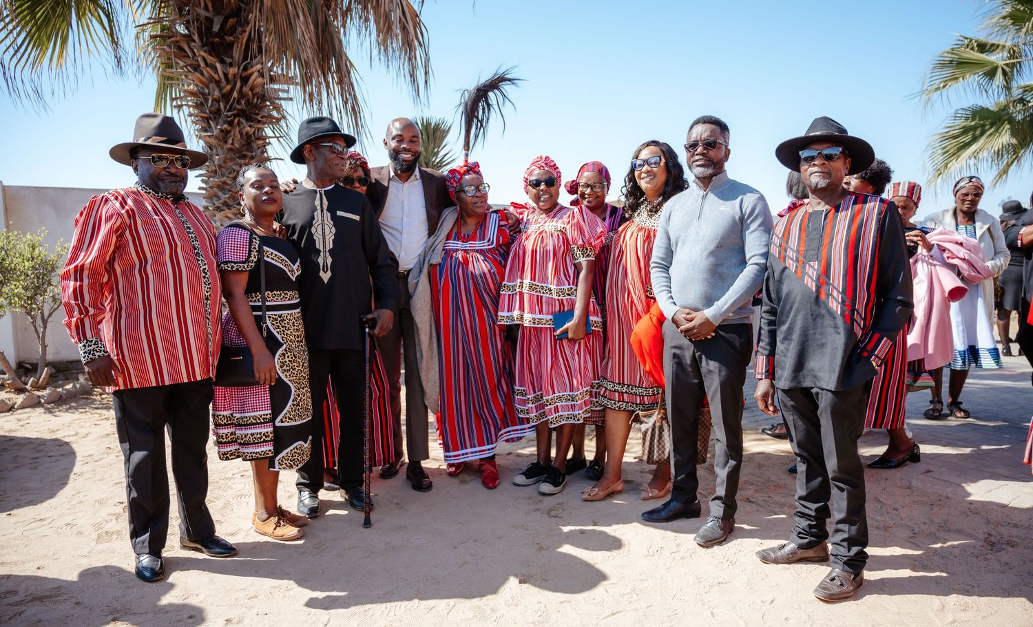 Group of people in vibrant traditional African attire posing together outdoors in a sunny setting.