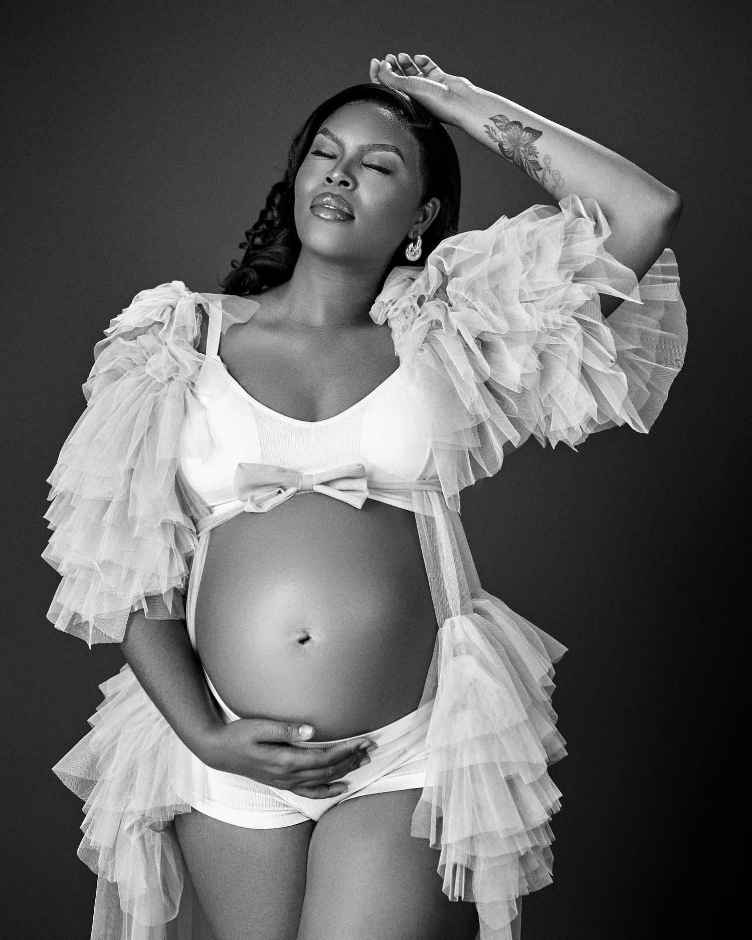 A black and white maternity photo featuring a woman in a ruffled white gown posing elegantly with her baby bump.