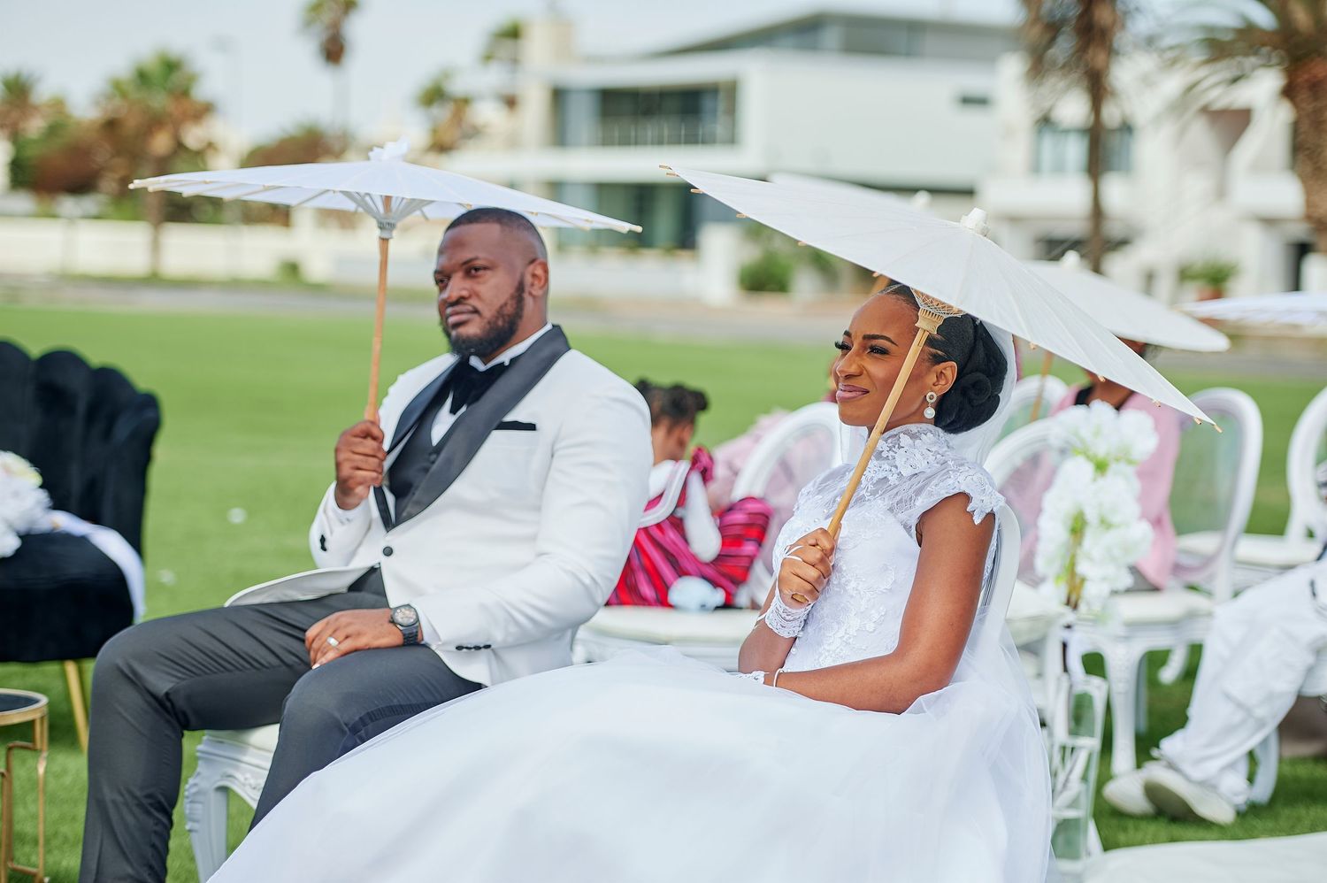Bride and groom sitting outdoors at wedding ceremony holding white parasols, dressed in elegant white attire on a sunny day.